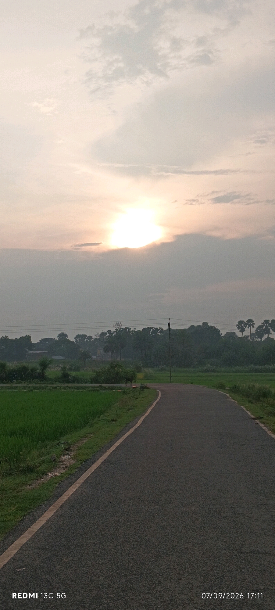 This is a vertical, outdoor, eye-level photograph taken at dusk, featuring a rural road winding through lush green fields and trees under a cloudy sky with the sun partially obscured by clouds. The overall scene suggests a tranquil setting in the Indian countryside.
The foreground is dominated by a paved, dark asphalt road with a single yellow line marking its edge. The road curves to the right, leading into the distance. To the left of the road, there is a vibrant green field, likely a crop of rice or similar vegetation. The ground adjacent to the field appears damp.
In the midground, a line of dense trees forms a dark silhouette against the sky. Some of these trees appear to be palm trees, indicated by their distinctive fronds. A solitary utility pole stands on the right side of the road, its top obscured by the trees. There are also some buildings visible behind the trees, suggesting a small village or settlement. A single cow is standing on the right side of the road, partially hidden by vegetation, facing away from the camera.
The sky is filled with clouds, with the sun shining brightly through a gap in the clouds in the upper center of the image. The light from the sun casts a soft, hazy glow over the scene, creating a peaceful and atmospheric effect characteristic of twilight.
The bottom left corner of the image displays a watermark: "REDMI 13C 5G". The bottom right corner shows a timestamp: "07/09/2026 17:11".
While the prompt mentions the "city of Barahat, India," this specific image does not contain any visual elements that directly identify it as Barahat, India. The scene is a generic representation of a rural Indian landscape.