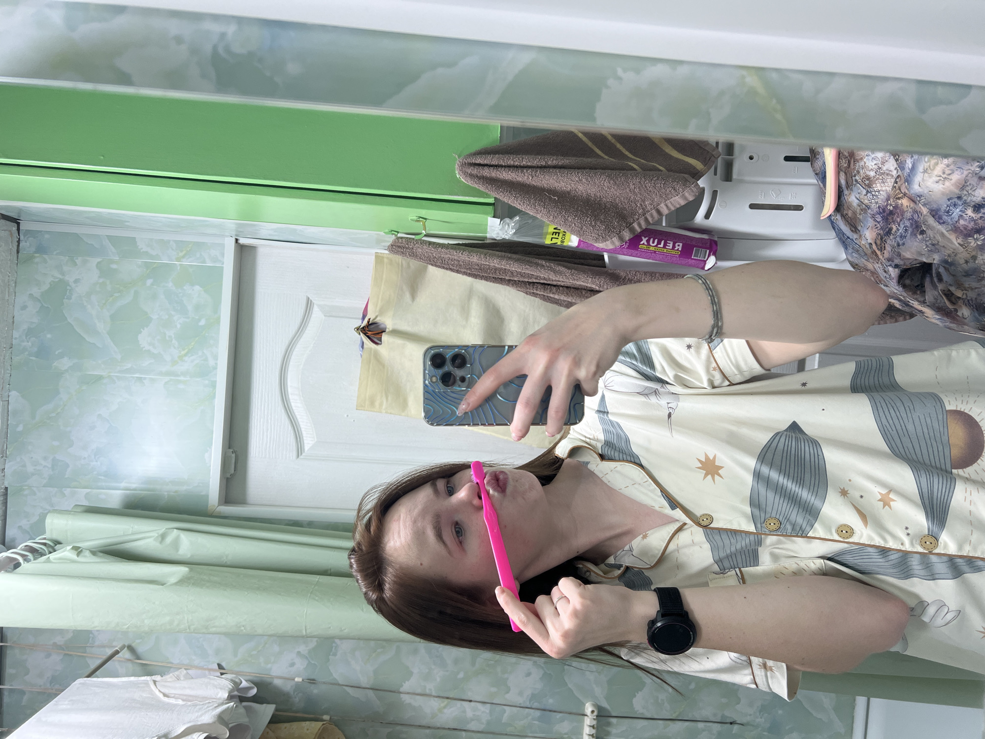 A young woman is taking a selfie in a bathroom. She is holding a pink toothbrush near her face, while also holding a smartphone. The bathroom has a green and white marble-patterned wall, a white door, and a mirrored medicine cabinet. There are towels and toiletries in the medicine cabinet. There is a black wristwatch on her left wrist. Her pajamas have a print of leaves and celestial elements. The location is presumed to be in Chaikovsky, Russia.