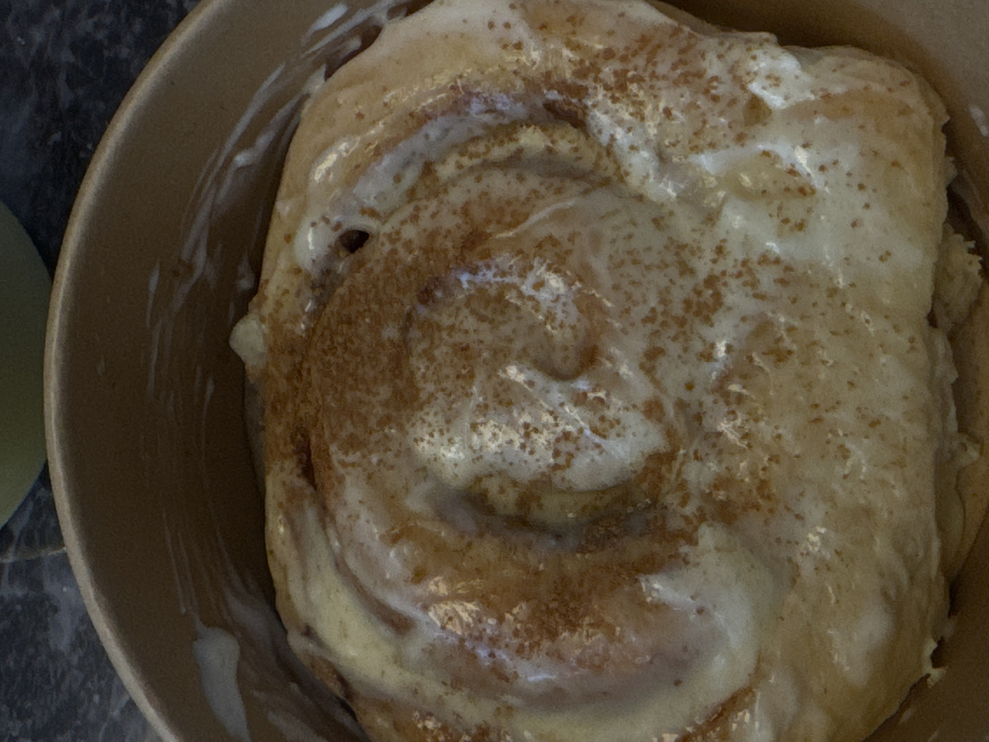 A close-up, overhead shot shows a cinnamon roll in a brown bowl. The roll is covered in white icing and sprinkled with brown spices. The bowl appears to be made of cardboard. Part of a green object is visible in the bottom left corner of the frame. The location is Unknown, Unknown.