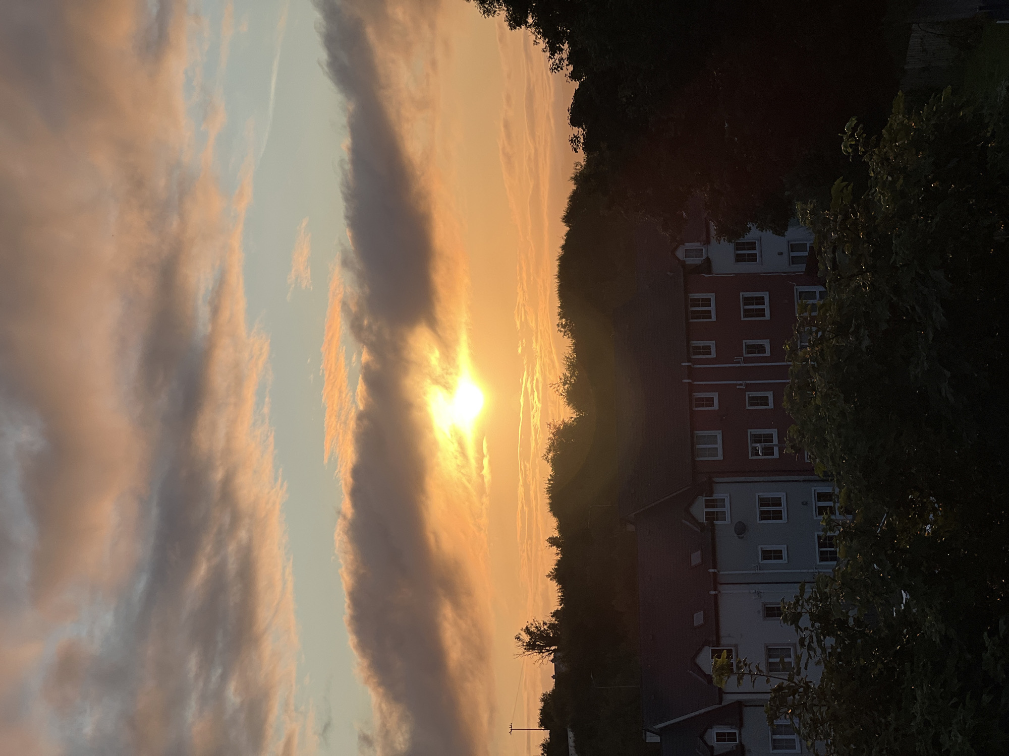 The image depicts a sunset over a residential area in Mountcharles, Ireland. The sky is filled with clouds tinged with orange and pink hues, illuminated by the setting sun. In the middle ground, a line of terraced houses with red and grey facades is visible, partially obscured by trees. The foreground consists of dense green foliage, also silhouetted against the sunset. The overall impression is one of a peaceful evening scene in a suburban or semi-rural setting. There are no people or discernible actions in the image.