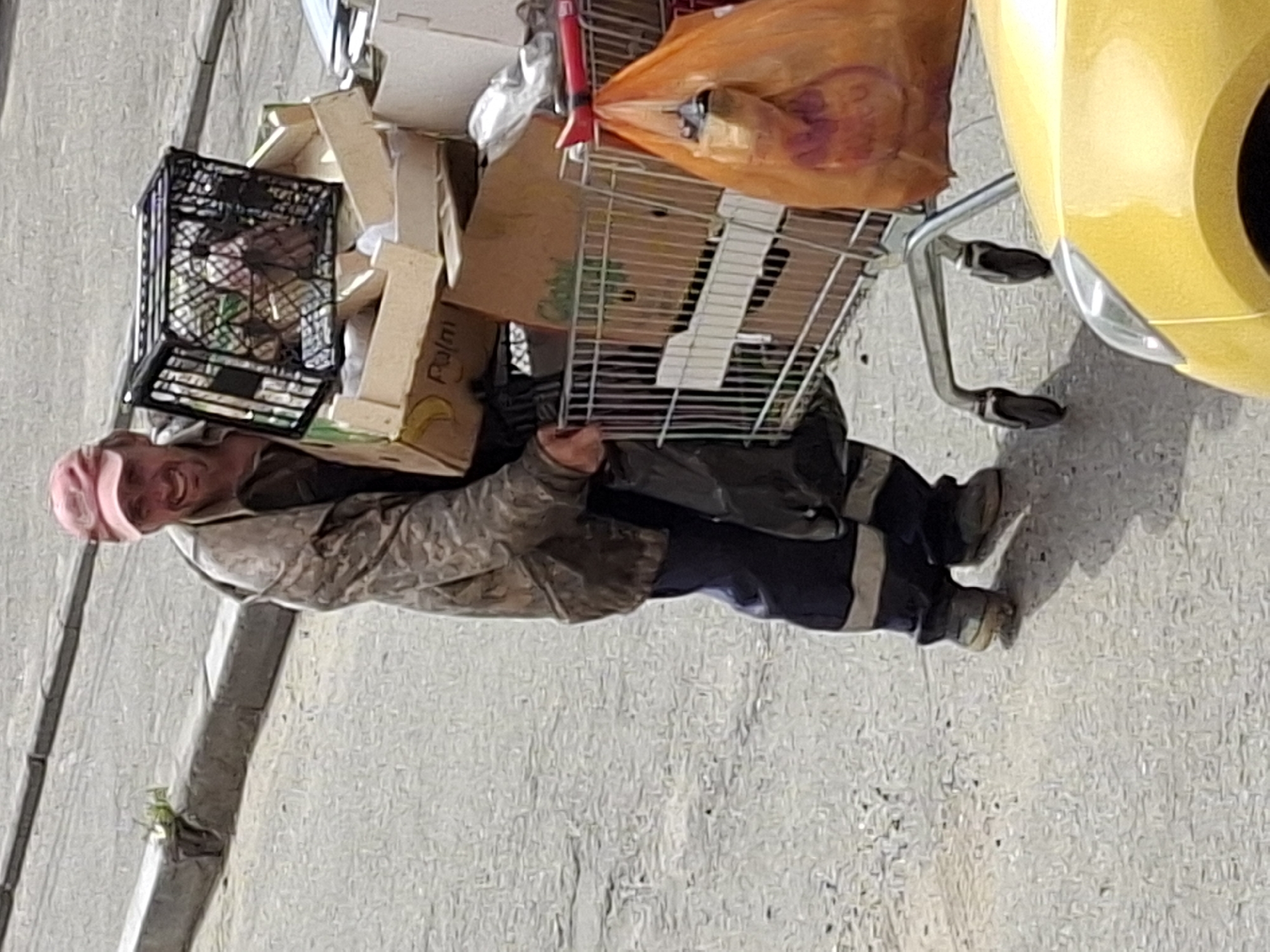 A smiling man wearing a pink cap and a camouflage jacket pushes a shopping cart filled with various items including boxes, plastic crates, and an orange plastic bag. He is walking on a paved street next to a curb with some weeds growing. Part of a yellow car is visible in the foreground.