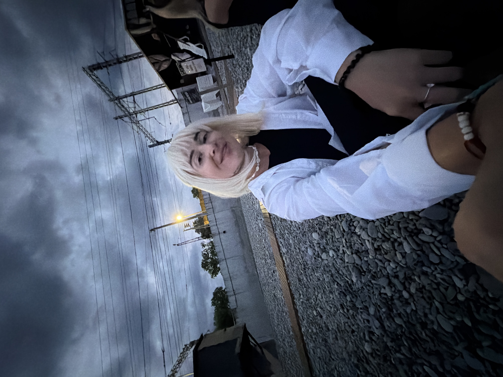 A medium shot, low-angle, natural light photograph shows a woman with short, blonde, choppy hair and bangs, wearing a white button-down shirt over a black top, and black pants. She is seated outdoors on a gravel surface, possibly near train tracks, as indicated by overhead wires and a distant train signal light. The woman is smiling and looking towards the camera, with her left hand resting on her lap and her right hand partially visible. She is wearing a silver ring on her right index finger and a beaded bracelet on her left wrist. The background features a concrete wall, trees, and overhead electrical infrastructure. The sky is cloudy and dark, suggesting either dusk or an approaching storm. The setting appears to be an industrial or transportation area, potentially on the outskirts of Sochi, Russia, given the presence of train infrastructure and a hint of coastal vegetation.