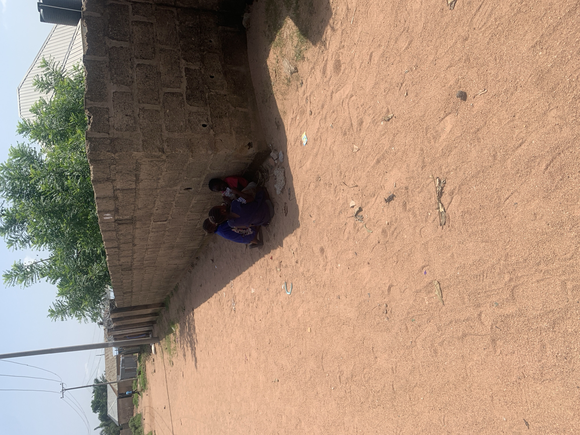 The image shows two children in Jalingo, Nigeria. One child, wearing a red shirt, is being held by an older child or adult wearing a blue garment. They are positioned in a shaded alcove formed by a brick wall and what appears to be the corner of a building. The ground is sandy and dry, with some scattered debris. A shadow from the wall stretches across the sandy area. Behind the wall, green foliage from a tree is visible. To the left, a portion of a corrugated metal roof and a water tank can be seen, suggesting this is an outdoor, possibly residential or communal space. The lighting indicates it is daytime, with the sun casting distinct shadows. The overall impression is one of a simple, outdoor scene in a warm climate.