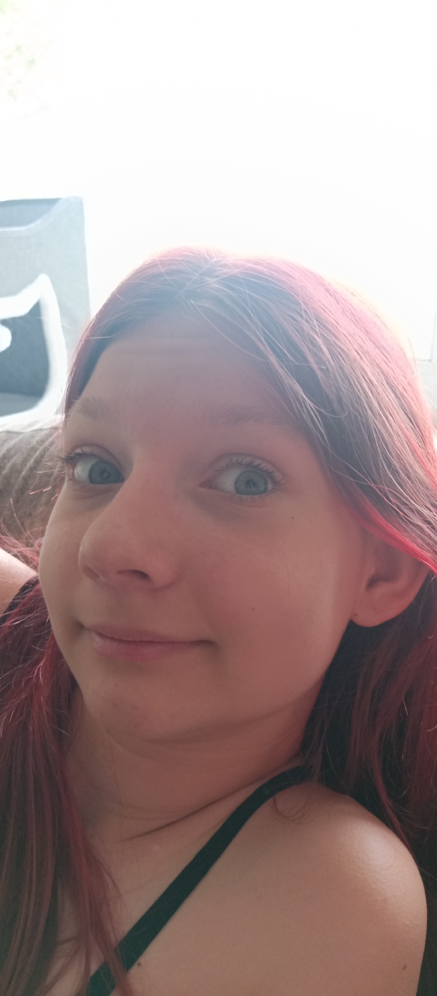 The image is a close-up of a person's face. The person has light skin, blue eyes, and red hair. The person's expression is slightly smiling. The person is wearing a black tank top. The background is blurry and appears to be an indoor setting. A grey object is in the top left of the photo. The city is unknown.