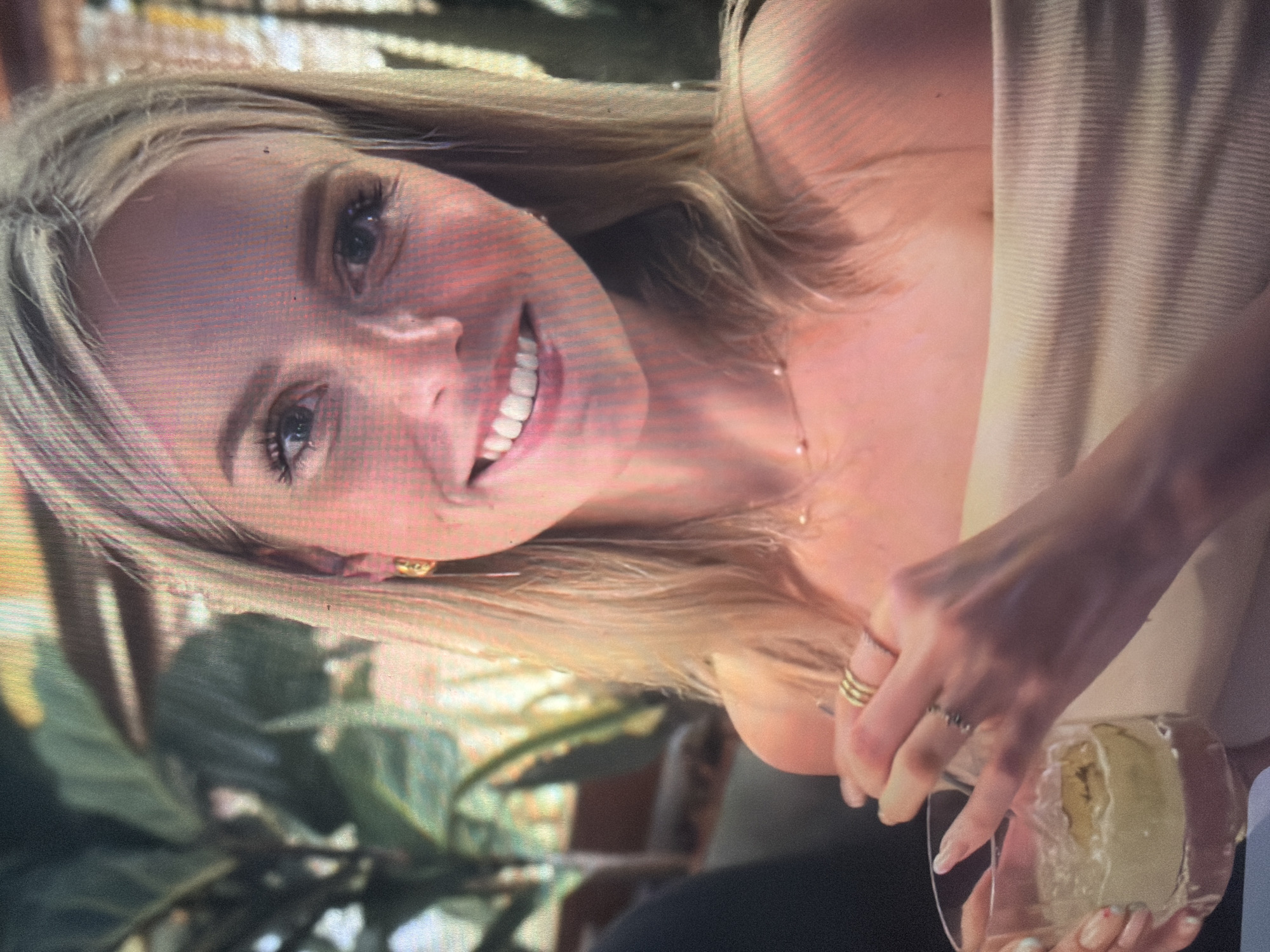 The image features a young Caucasian female smiling directly at the viewer, seated in what appears to be a relaxed social setting in the city of Shumen, Bulgaria. She has shoulder-length blonde hair, blue eyes, and visible eyelashes. Her skin is light-toned, with subtle freckles across her nose and cheeks. She wears gold hoop earrings and a delicate gold chain necklace adorned with small pearls. Her attire consists of a light beige, ribbed off-the-shoulder top. Her right hand, decorated with multiple gold rings (three stacked on her ring finger and another on her middle finger), holds a clear glass containing an iced beverage with a light-colored liquid and a visible garnish, possibly a citrus peel or wedge. The background is softly blurred, showing lush green foliage, suggesting an indoor garden or patio environment, with hints of warm-toned structural elements.