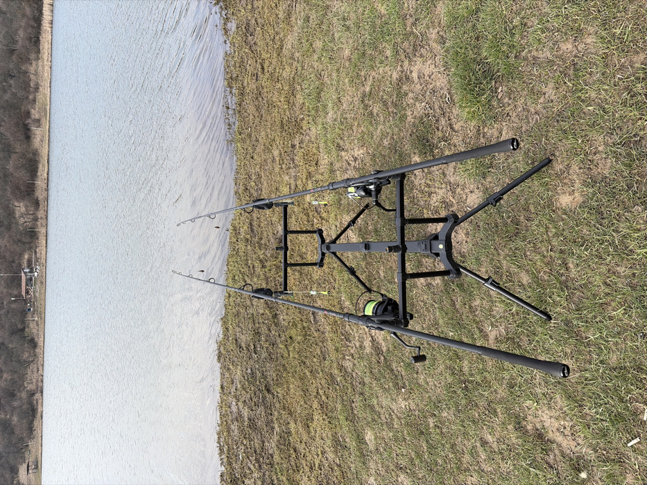 The scene is a lakeside fishing setup in Cherna Gora, Bulgaria. In the immediate foreground, a black, three-legged fishing rod pod supports two dark fishing rods, positioned horizontally and angled towards the water. Both rods are equipped with reels spooled with yellow-green fishing line, and bite indicators featuring yellow-green elements are suspended from the lines. The ground beneath the equipment consists of sparse, dry grass and patches of exposed earth. The midground features a calm body of water with minimal surface ripples. On the distant bank, a line of bare deciduous trees stretches horizontally, partially obscuring a small, light-colored building with a dark roof, likely a hut, visible towards the left. No individuals are visible within the frame.