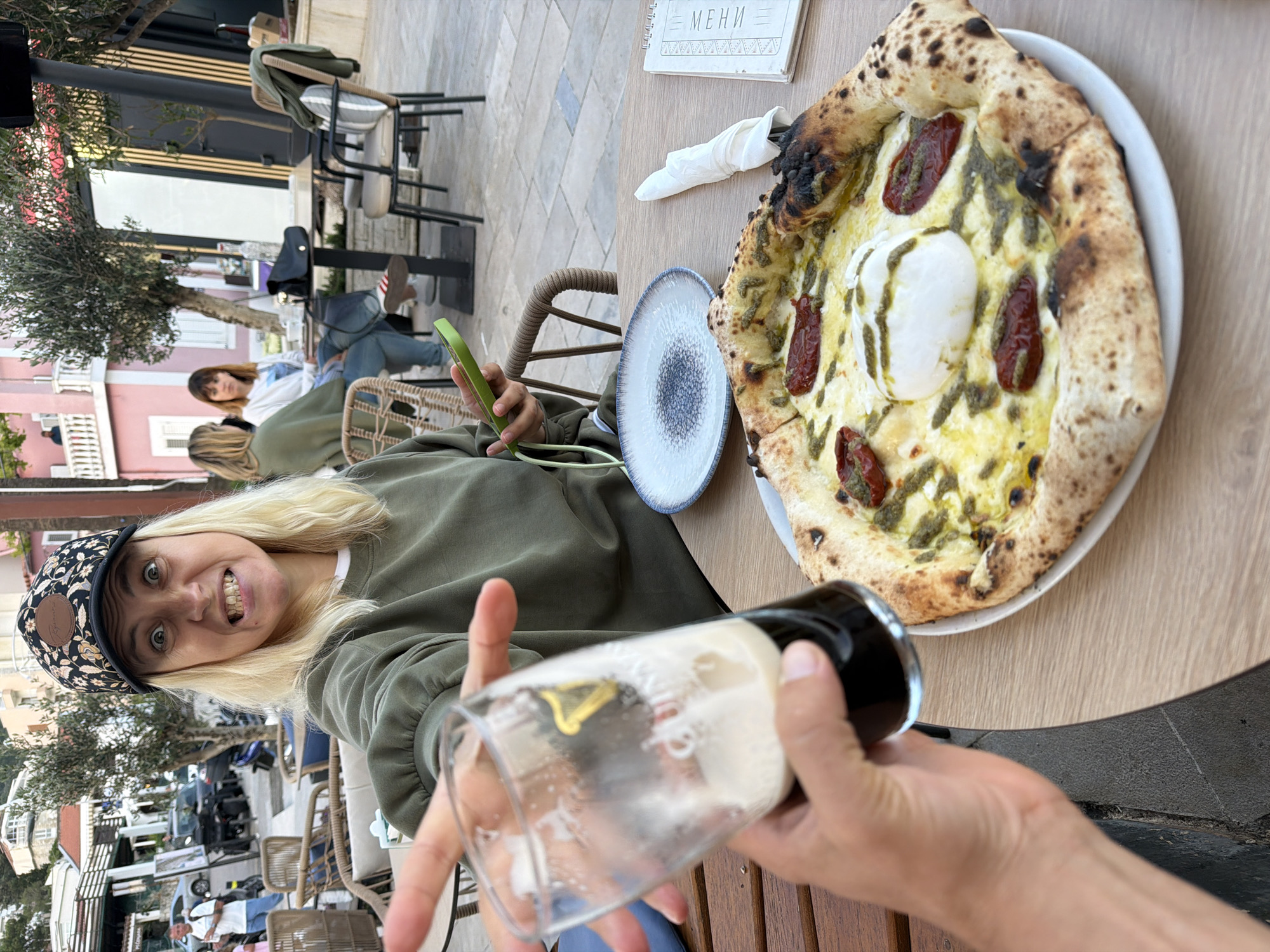 The image, likely taken in Petrovac centar, Montenegro, shows a woman wearing a floral cap and green sweater, positioned at an outdoor table. She smiles at the camera, while holding a glass of dark beer. At the table is a pizza with what appears to be burrata, pesto, and roasted tomatoes. Next to the pizza is a small plate, a napkin, and a menu. In the background there are other people seated at tables and buildings, including a pink structure with a white balcony. Some vegetation is visible.