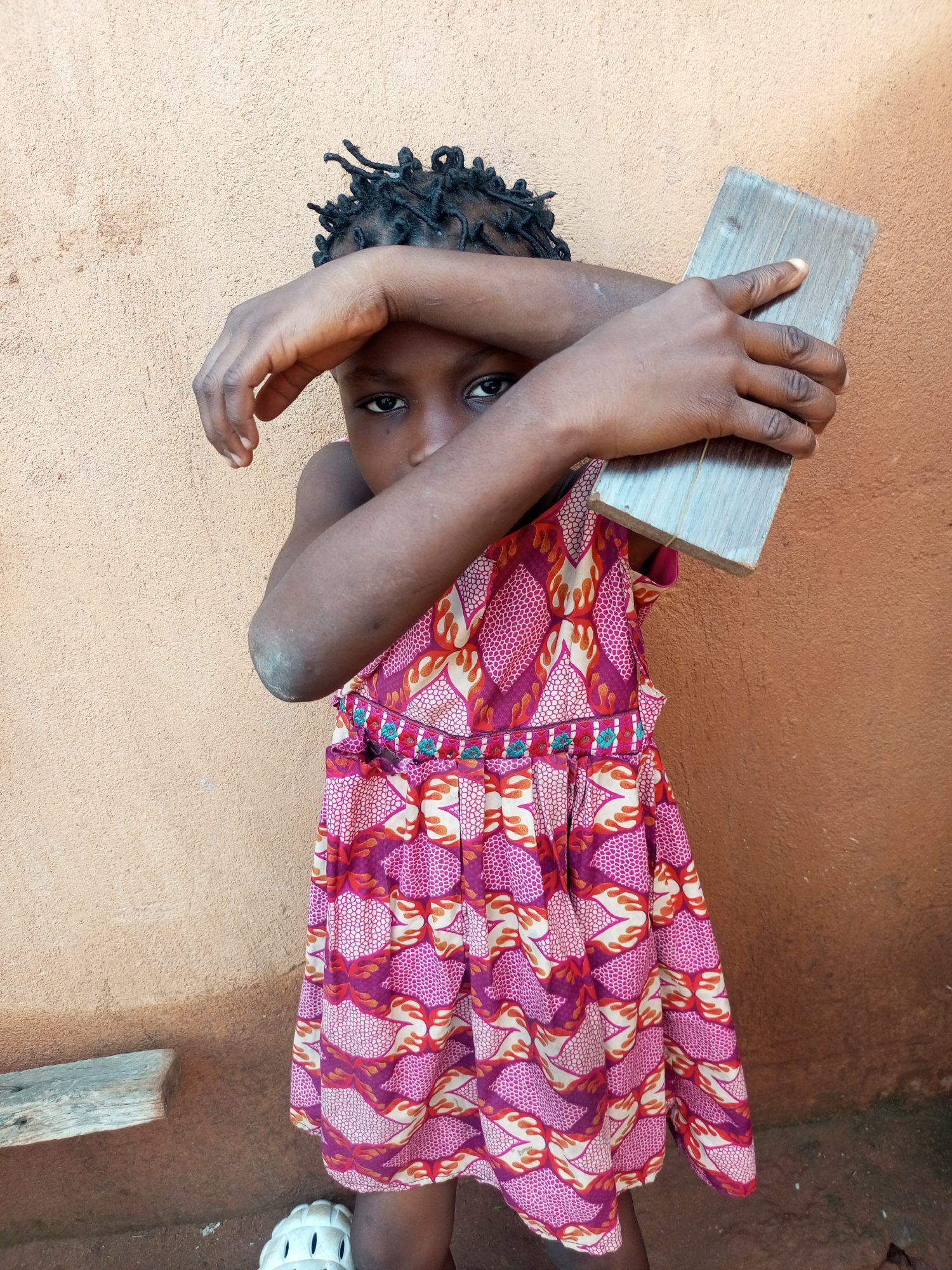 A young girl with dark skin and braided hair stands against a textured, peach-colored wall. She is wearing a pink, purple, and white patterned dress. Her arms are crossed in front of her face, and she is holding a light-colored wooden plank. Her knees are visible, and she is wearing white sandals. The ground is composed of dirt and some small rocks. To the left, a weathered wooden plank is horizontally placed against the wall. The scene appears to be outdoors, possibly in a rural or less urbanized area of Tempure, Nigeria.