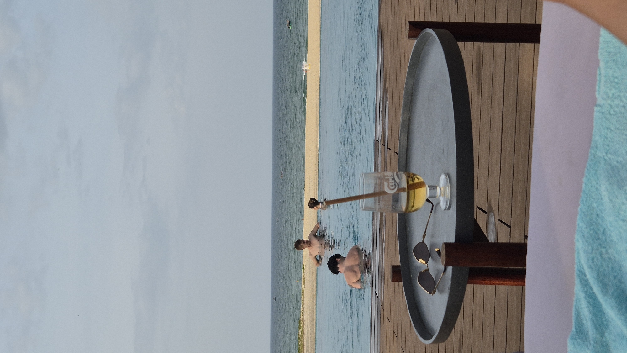 The image depicts an infinity pool overlooking the ocean, with a sandy beach and the sea in the background. In the foreground, a round, dark-grey table with wooden legs is set on a wooden deck. On the table, there are a pair of sunglasses and a glass of amber liquid with a straw. The glass has the "Carlsberg" logo visible. In the pool, three people are visible; two men and one woman. One man is facing away from the camera, his back to the viewer, and is standing in the water. Another man, facing the camera, is also standing in the water, with his hands on the edge of the pool. A woman with dark hair is standing in the water to the right of the man facing the camera. The sky is overcast. The overall scene suggests a resort or hotel setting in Hoi An, Vietnam, likely along the coast.