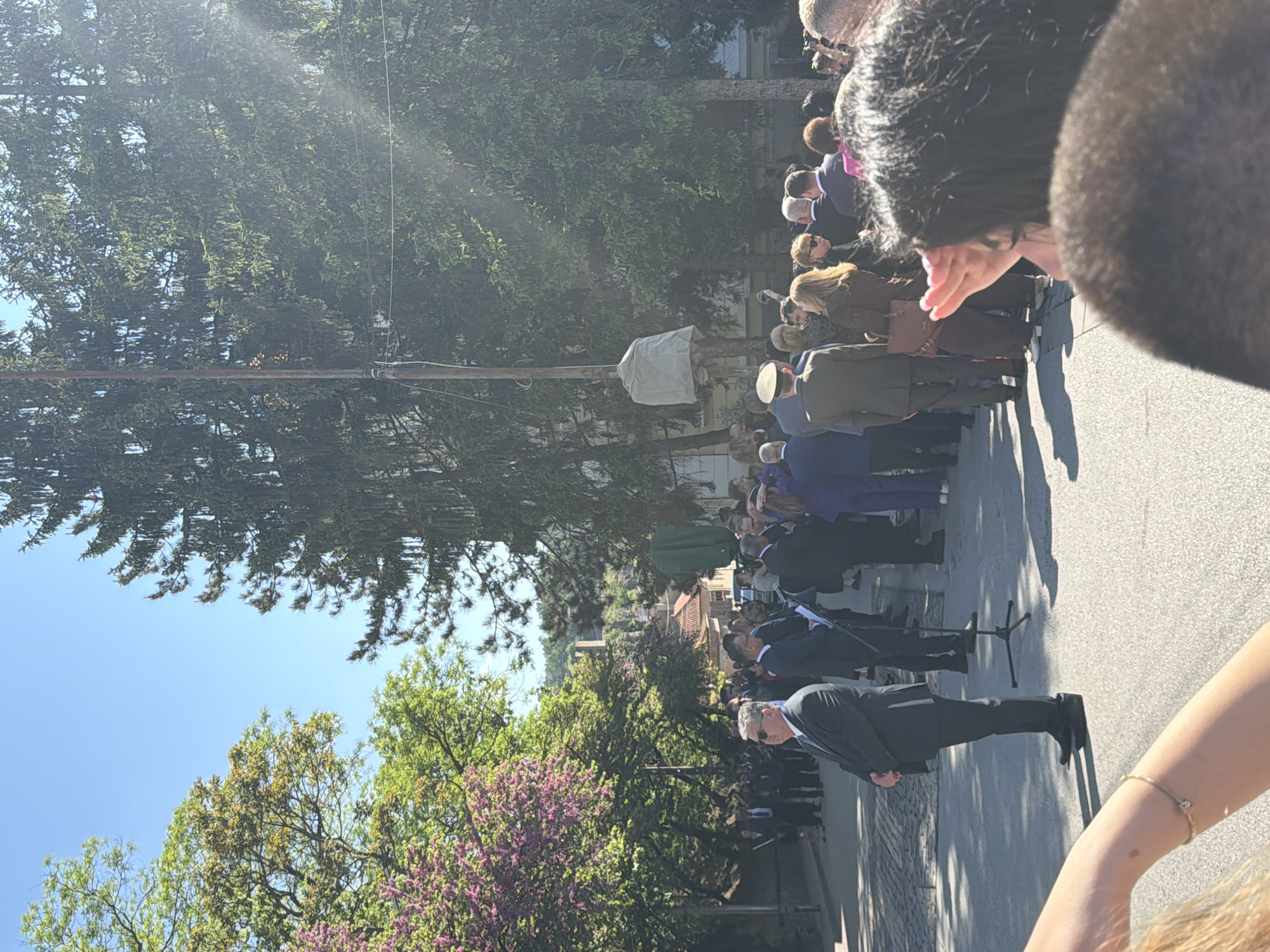 The image shows a gathering of people standing on a street in Veliko Tarnovo, Bulgaria. Most individuals are dressed in formal attire. One man is standing at a microphone addressing the crowd. Trees line the side of the street. The sky is blue and the weather appears sunny. There are older buildings in the background.