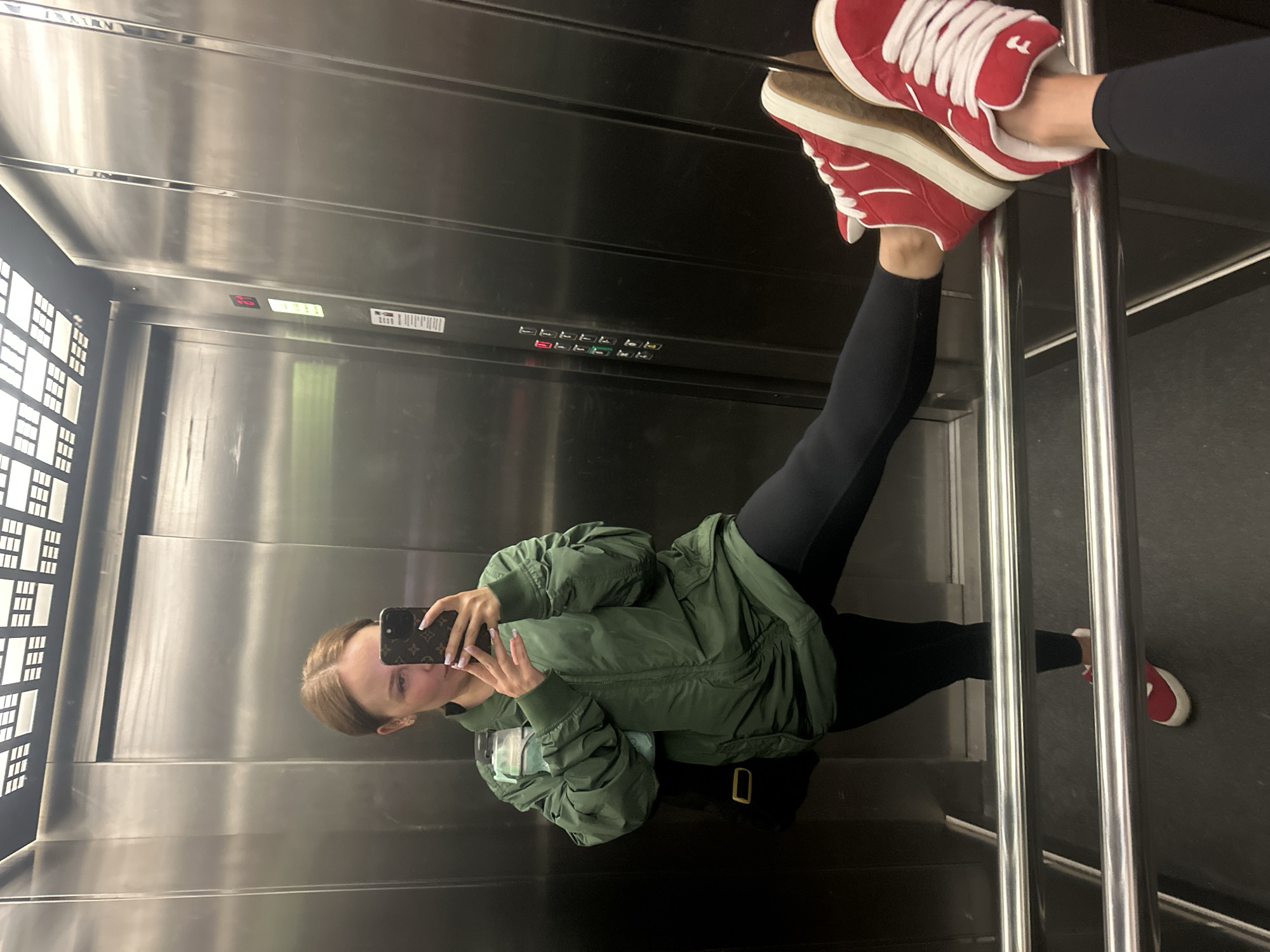 A person is taking a selfie in an elevator. The elevator has a steel interior with visible buttons and a window. The person is wearing black leggings, a green jacket, and red sneakers with white laces. The selfie captures the person holding a phone in front of them, reflecting their image in the elevator's steel wall. The setting is an elevator; the city of Budva, Montenegro, is not discernible from the image.