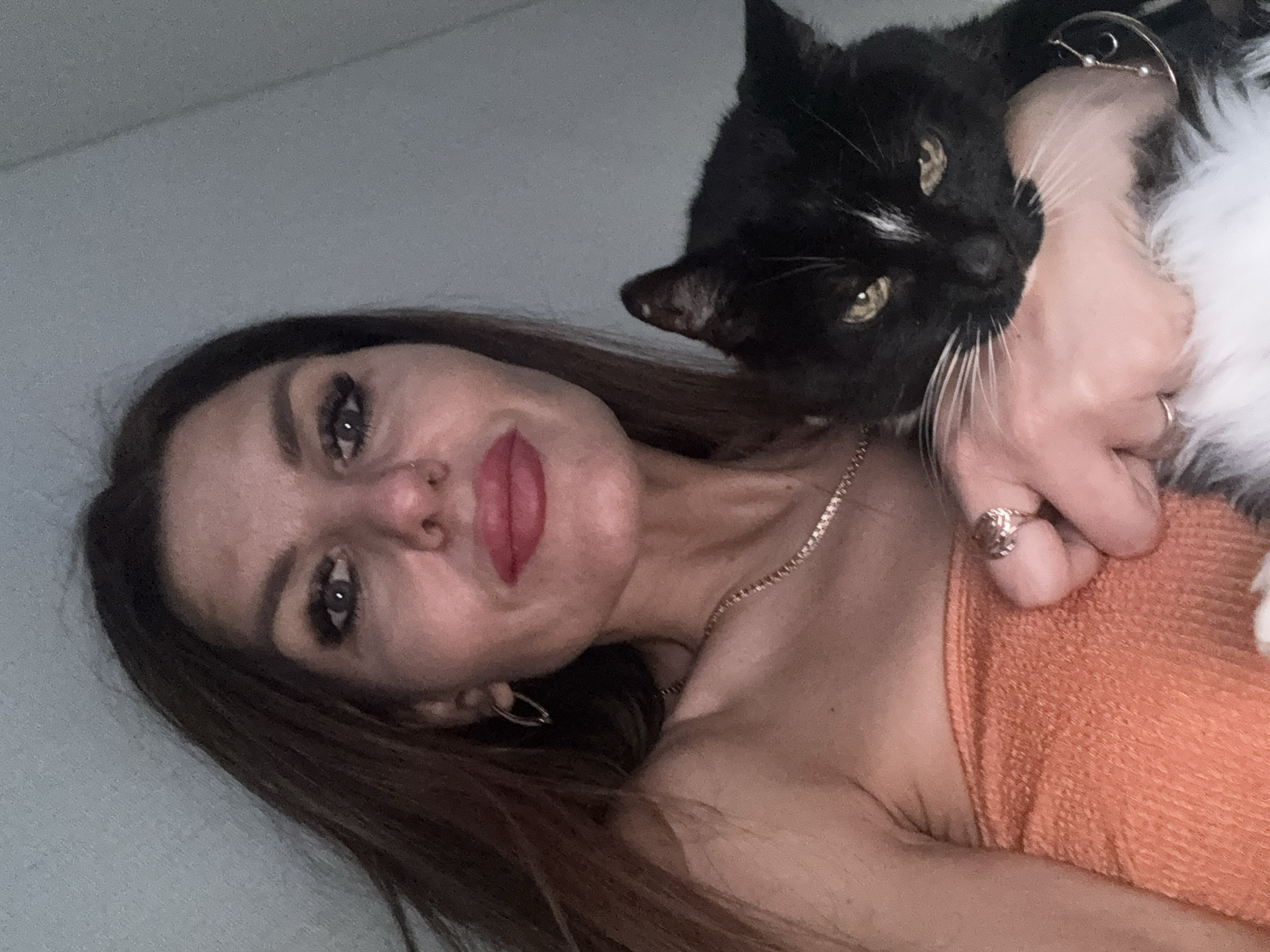 A woman with long brown hair and fair skin is holding a black and white cat. She is wearing an orange strapless garment and a gold chain necklace. She has a nose ring and earrings. The background is a light blue wall. The location in the city of Chaikovsky, Russia is not specified in the image.