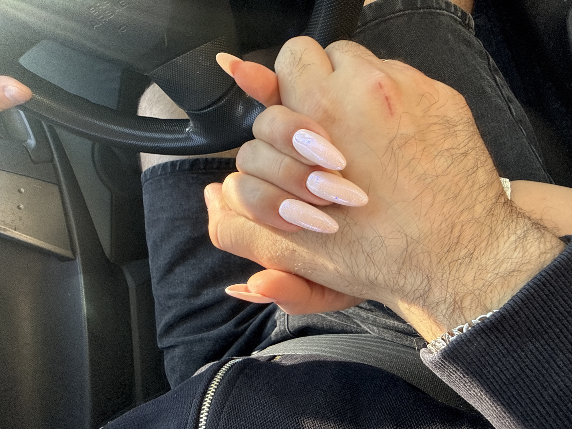 The image shows a close-up view of two people holding hands inside a car. One person has long, almond-shaped nails painted with a pearlescent pale pink polish. Their hand is on top of the other person's hand, which has visible hair and a visible scrape on the back. Both individuals are wearing dark denim jeans. The interior of the car, including the dashboard and steering wheel, is visible in the background. The lighting suggests it is daytime, with sunlight illuminating parts of the scene. There is no visible indication of the location being in Tallinn, Estonia.