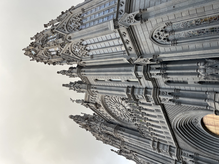The image shows the exterior of a large, gothic-style church building in Arucas, Spain. The church is constructed of gray stone and features a large rose window, a central spire, and numerous other ornate details. The building is a prominent landmark in the city and is located at 28.119 latitude, -15.523 longitude. It appears to be a cloudy day. The church is an architectural marvel and is a testament to the skill of the builders. The church is the Iglesia de San Juan Bautista, which is one of the most important churches in Arucas. The church is a popular tourist destination, and visitors can admire its intricate architecture and learn about its history.