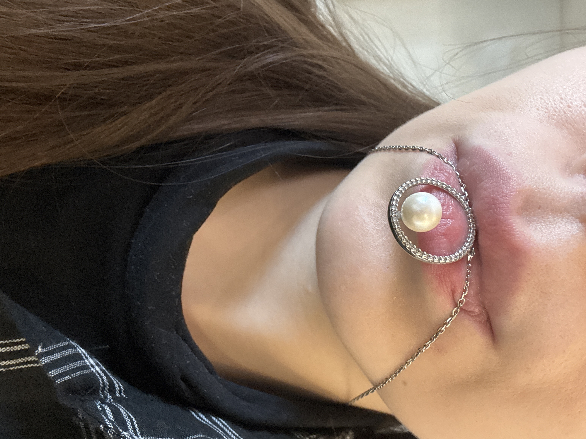 The image is a close-up, overhead shot of a person's chest and neck area, with their hair in the upper left corner. The person is wearing a black top, with a portion of a black and white striped fabric visible at the bottom. A silver chain with a circular pendant featuring a pearl and clear stones is draped over the person's décolletage. The lighting is neutral, suggesting an indoor setting. No specific time of day, weather, or location cues are discernible beyond the provided context of Ladozhskoe Ozero, Russia. No text is visible in the image.