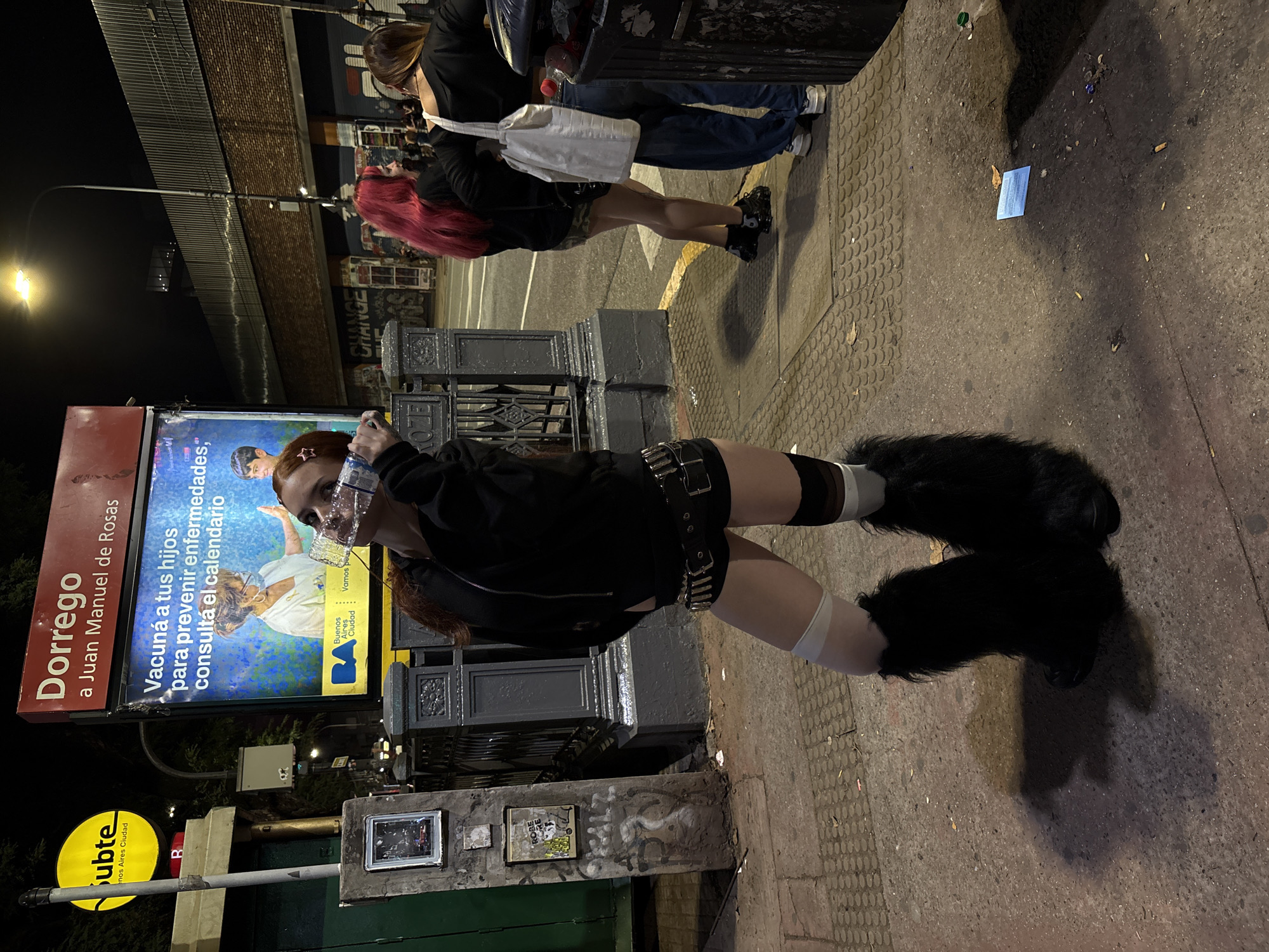 The image shows a street scene in Buenos Aires, Argentina, at night. A person wearing a black jacket, shorts, a studded belt, and large, furry black boots stands on a sidewalk. They are wearing a plastic bottle over their face. Behind them is a public transportation sign for the "Subte" (subway) with "Dorrego a Juan Manuel de Rosas" written on it. The sign also includes a health message: "Vacuná a tus hijos para prevenir enfermedades, consultá el calendario." A poster of the "Subte" is yellow. Other people are walking by on the sidewalk in the background, some with red hair. There is graffiti and artwork on nearby surfaces. The street is paved and has some litter. There is a lamp post shining light on the street.