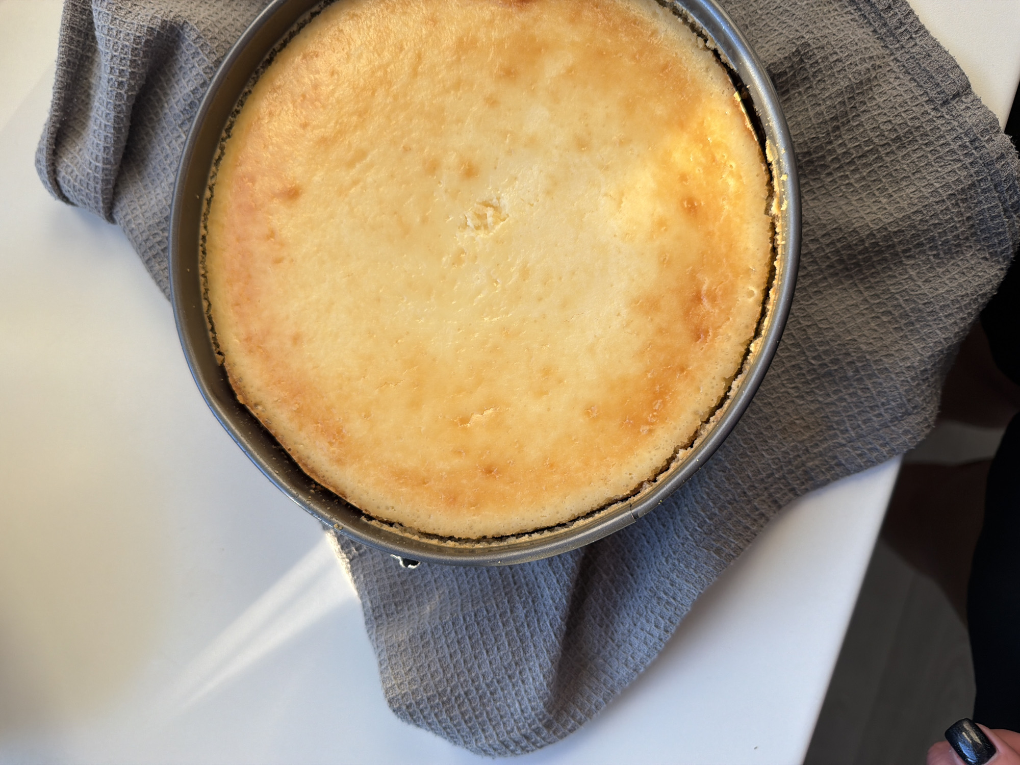 A baked cheesecake rests in a springform pan on a gray waffle-weave dish towel. The cheesecake is golden brown on top with slight surface imperfections. The pan is metallic gray. The towel partially covers a white surface. In the lower right corner, a small portion of a person's hand with a dark fingernail is visible. The location is not discernible from the image.