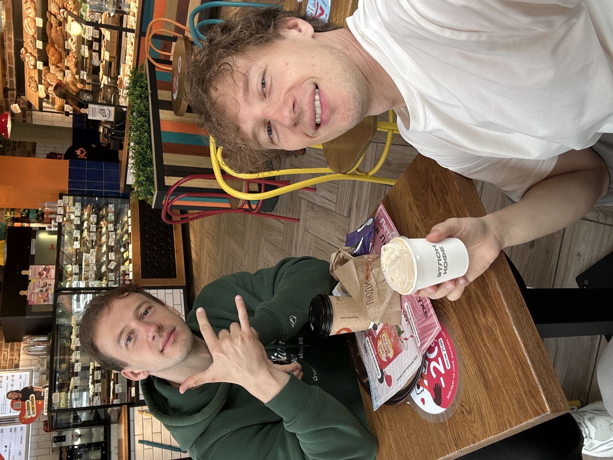 Two men are seated at a wooden table in a cafe. The man on the left, wearing a dark green hoodie, gestures with his hands. The man on the right, wearing a white t-shirt, smiles at the camera and holds a white paper cup with a lid. On the table are a brown paper bag, a black and brown disposable coffee cup, and various papers with Cyrillic text and images. Behind the men, display cases filled with pastries and bread are visible, along with colorful chairs and brick walls, suggesting a bakery or cafe setting. The location is identified as Murino, Russia.