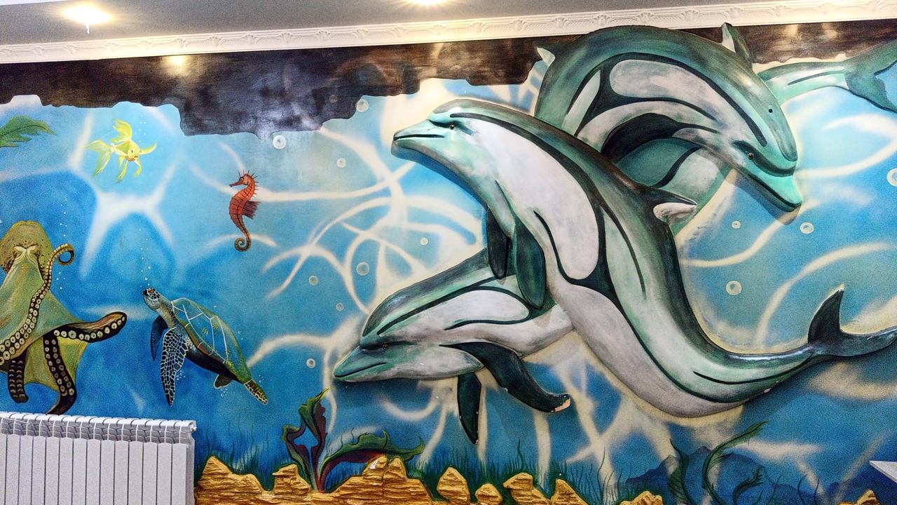 The image depicts a vibrant mural on a wall, likely in an interior space, showcasing an underwater scene. The mural features several marine creatures rendered in a three-dimensional, sculpted style. Prominently displayed are three dolphins, two of which are larger and in the foreground, appearing to swim dynamically. Their bodies are a mix of light gray and teal, with dark outlines. Behind them, a third, smaller dolphin is partially visible. To the left of the dolphins, a large, reddish-brown octopus with visible suckers on its tentacles is depicted. Adjacent to the octopus, a green sea turtle with intricate patterns on its shell is swimming. Further to the right and higher up, a small, bright orange seahorse curls its tail. In the upper left quadrant of the mural, a small, yellow fish with flowing fins is present. The background is a gradient of blue, simulating water, with abstract white swirls and bubbles suggesting movement and light refraction. The bottom of the mural transitions into a rocky, textured base with green seaweed. On the far left, a white ribbed radiator is partially visible at the bottom of the frame. The top of the image shows a white decorative ceiling border and a light fixture. The artwork is detailed and colorful, creating an immersive ocean environment. The scene is static, with no people or overt actions taking place. The location in the city of Balykchy, Kyrgyzstan, is not visually ascertainable from the image itself.