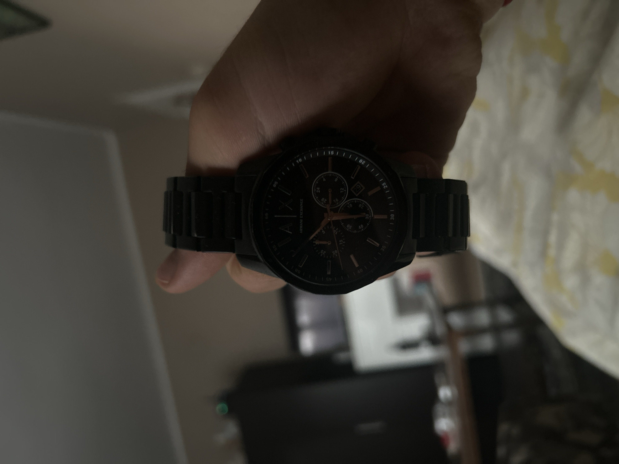Here is a forensic description of the image:
A person's hand holds a black Armani Exchange wristwatch. The watch has a black face with rose gold-colored hands and numerals. The watchband is also black and appears to be made of metal links. The background is blurry, but it seems to show a room with a white wall and a piece of furniture. The environment is indoors and the lighting is slightly dim.