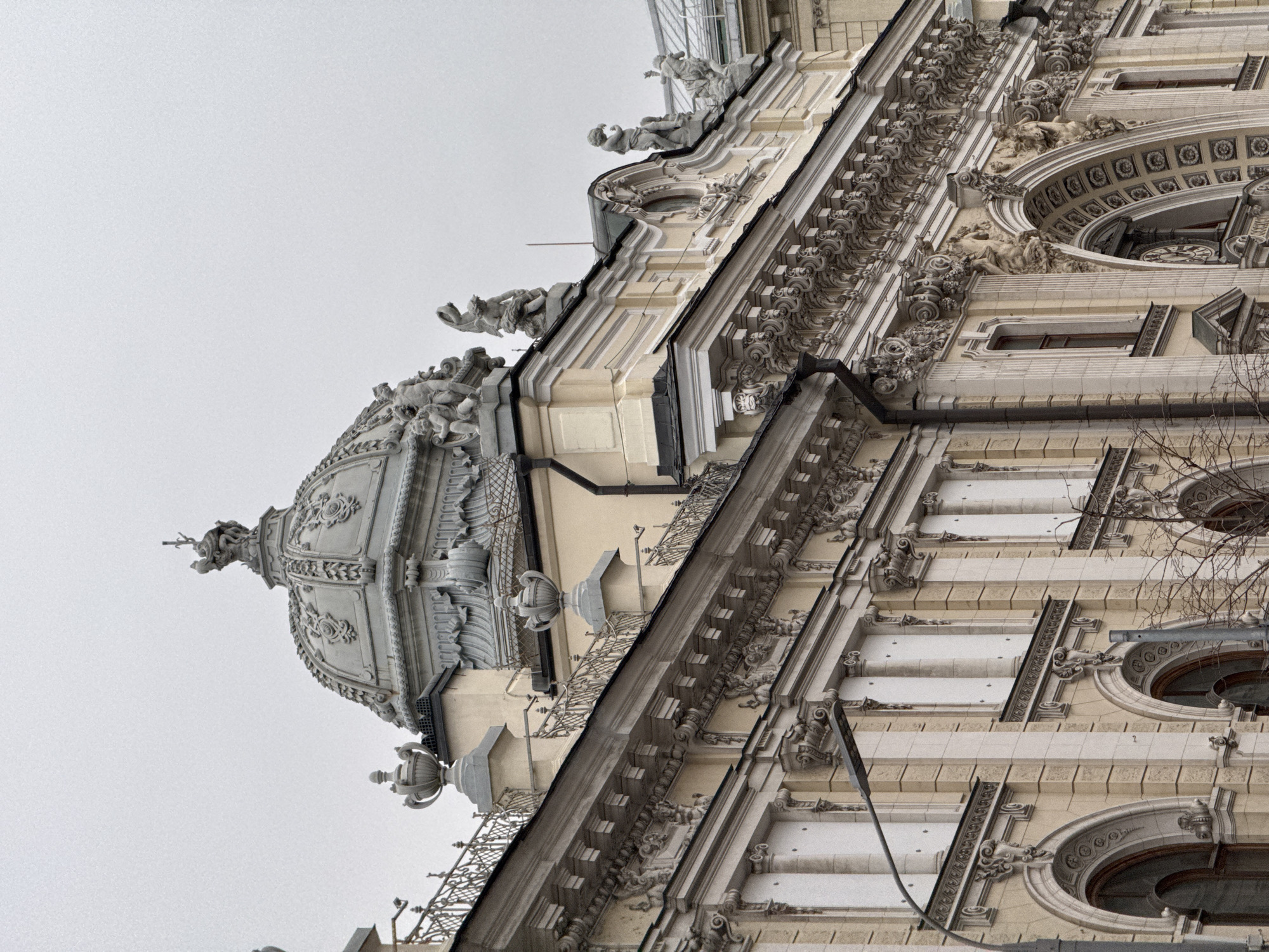 The image shows the architectural details of a building in Moscow, Russia. The building has a decorative dome topped with a spire. There are ornate carvings, statues, and rounded windows. The overall architectural style is Baroque. The sky is overcast.