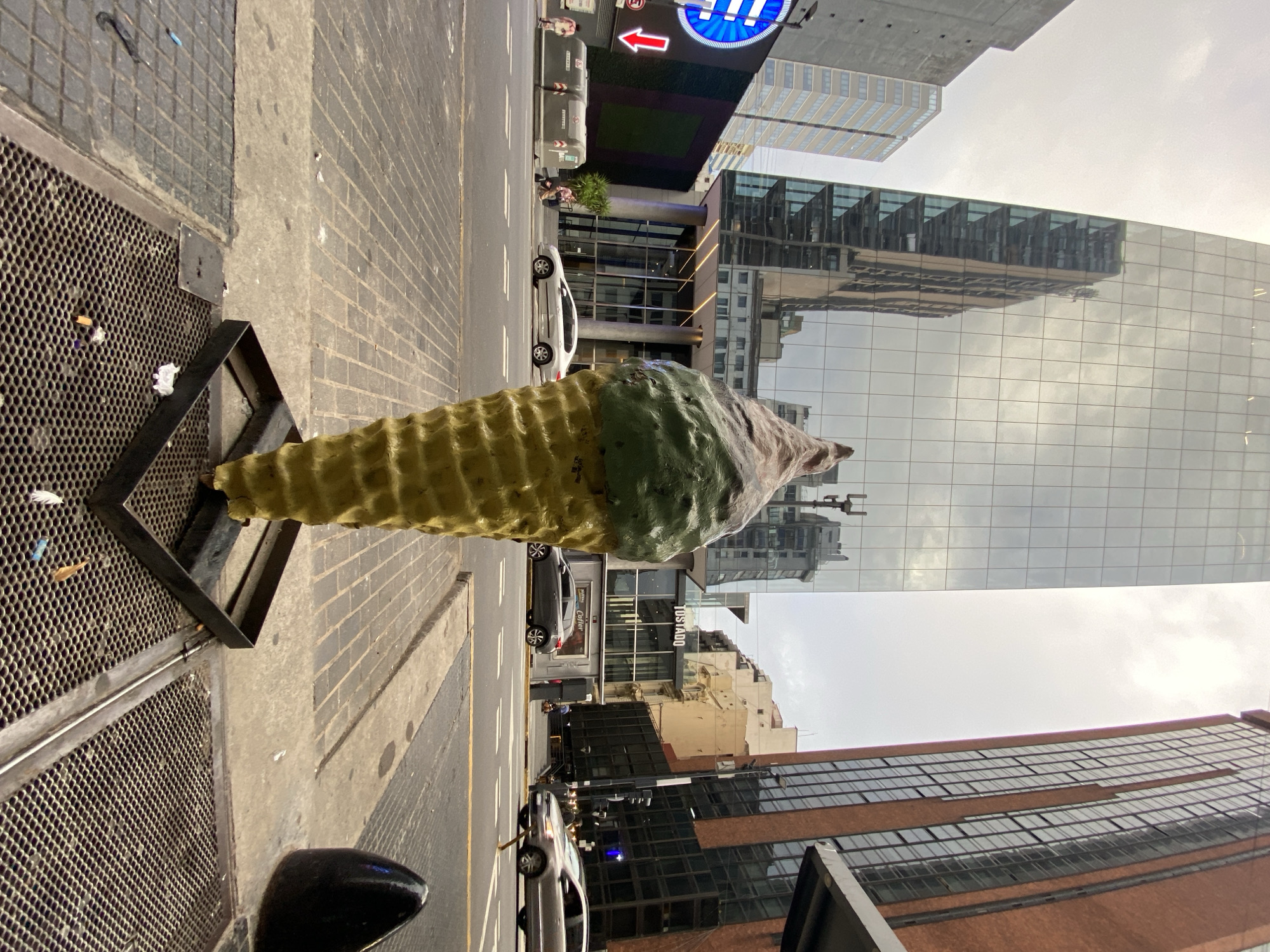 The image depicts a low-angle, street-level view on Avenida Corrientes in the Microcentro neighborhood of Buenos Aires, Argentina, under an overcast sky. A large, weathered sculpture of an ice cream cone stands centrally in the foreground, approximately 2 meters tall. The sculpture features a yellow textured cone base, topped with two scoops of ice cream, one green and one brown/beige. It rests on a black square metal base, situated over a metal grate covering part of the sidewalk. Scattered debris, including small pieces of paper and cigarette butts, is visible on and around the grate.
The midground features a multi-lane asphalt street with white lane markings and yellow crosswalk lines. A silver sedan is parked parallel to the curb across the street, in front of a building entrance. Further right, a grey SUV is partially visible, moving right to left.
In the background, a tall, modern glass skyscraper dominates the left side, reflecting the overcast sky and elements of surrounding buildings. Its lower section features a multi-story glass and columned entrance. To the right, a multi-story building with a red brick facade and numerous dark-framed windows is prominent. Below it, across the street, a commercial establishment with a visible "TOSTADO" sign is present. Pedestrians are scattered on both sidewalks: two individuals are visible on the left, near a large blue circular sign with a white downward arrow and a row of grey street bins. Other pedestrians are indistinctly visible on the far sidewalk, navigating the commercial environment. The scene portrays an active urban commercial district during daylight.