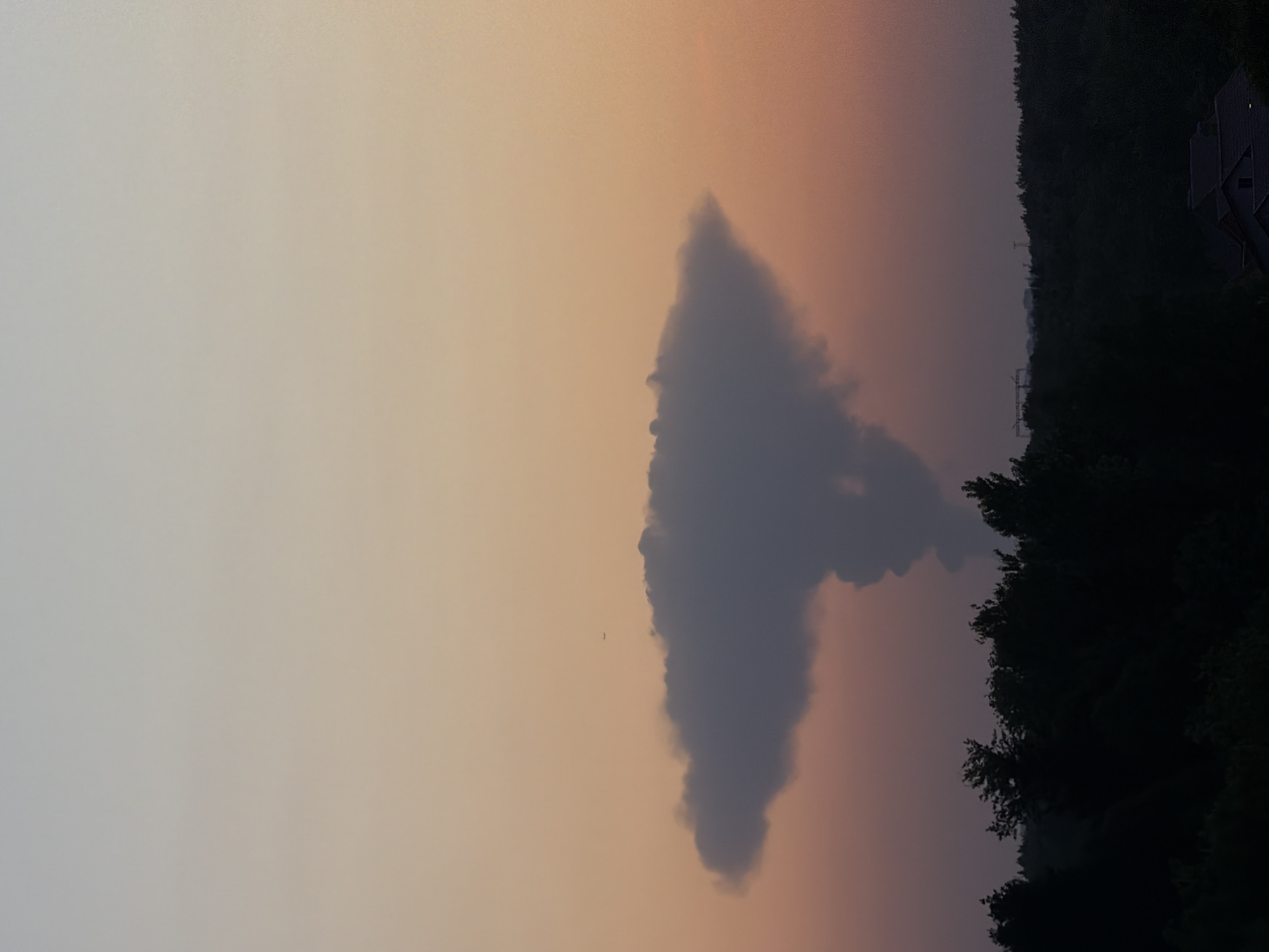 The image shows a landscape with a prominent cloud formation against a gradient sky transitioning from light gray to orange and pink hues. The cloud is dark gray and roughly triangular in shape, with a distinct peak at its top. To the right of the cloud is a dense treeline silhouetted against the sky, suggesting a forest or wooded area. A structure resembling a house or building is visible on the far right edge of the frame, nestled among the trees. Power lines are also visible near the treeline. A tiny flying object, potentially a bird, is located near the center cloud.