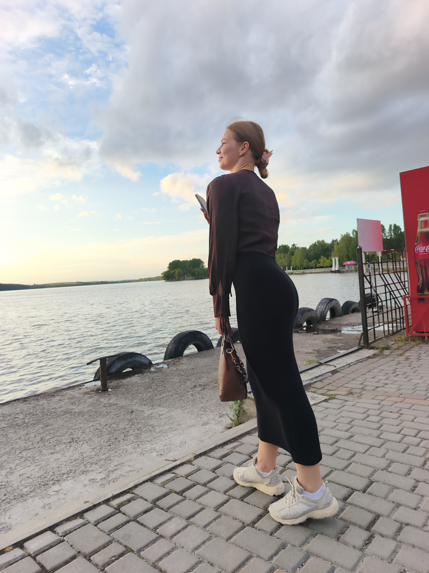 A woman stands on a paved walkway near a body of water, looking out at the horizon. She is wearing a dark brown long-sleeved shirt, a long black skirt, and light-colored athletic shoes. A brown handbag with a chain strap is held in her left hand. She is holding a smartphone in her right hand. In the background, there is a body of water, trees on the opposite shore, a metal fence, and a red Coca-Cola advertisement. The sky is partly cloudy with a mix of blue sky and grey clouds. The scene appears to be a public promenade or waterfront area in Ternopil, Ukraine.