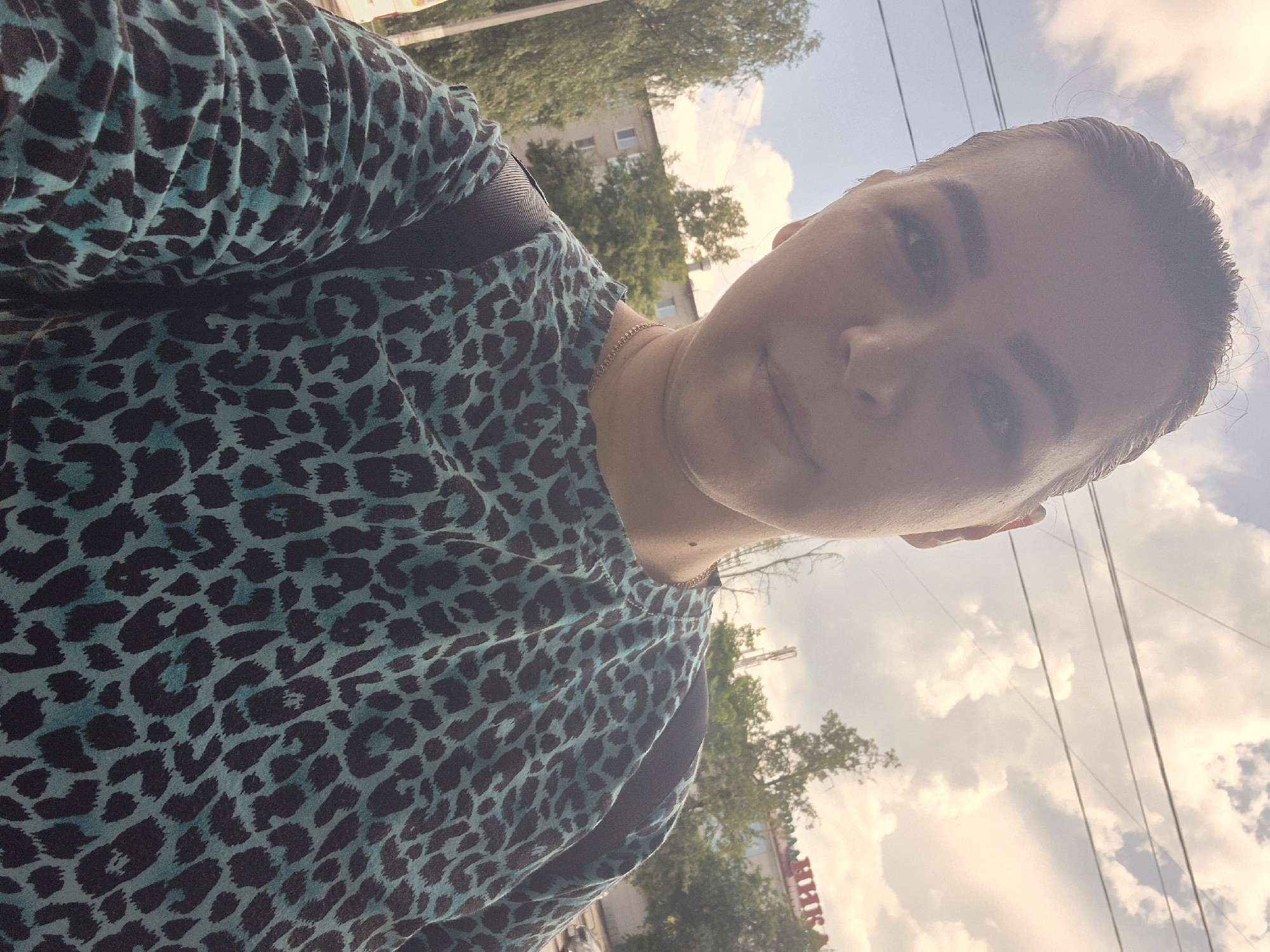 In a selfie taken outdoors in Chaikovsky, Russia, a young woman with her hair pulled back is the central subject, looking towards the camera. She is wearing a teal and black leopard-print shirt. Behind her, a multi-story apartment building is visible, partially obscured by trees. The sky is partly cloudy. The foreground shows part of the woman's upper body and shoulders, with a black backpack strap visible over her left shoulder. A thin chain necklace is visible around her neck. The image appears to be taken during daylight, with soft lighting.