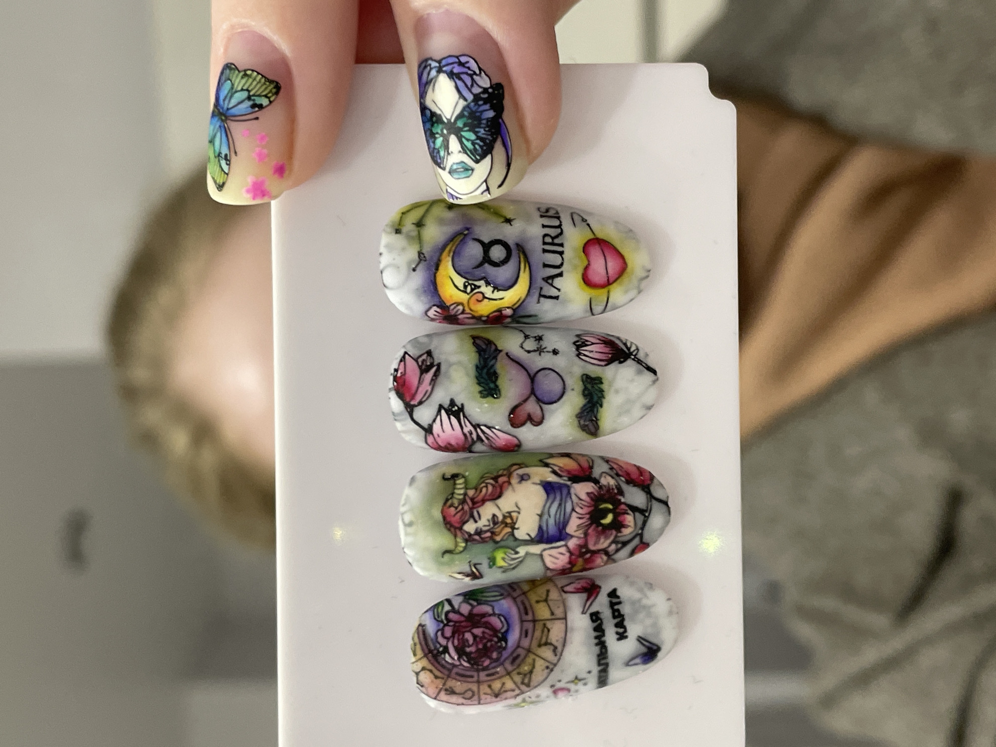 The image shows a hand holding a white rectangular surface displaying a series of five fingernail art designs. The fingernails holding the surface are painted; one with a butterfly and pink flower accents, and another featuring a woman's face with a butterfly mask. The designs on the white surface include astrological themes, such as zodiac signs, illustrations of figures, and floral patterns. The fifth nail includes Russian text that translates to "natal chart." The background is blurred, showing a person wearing a grey shirt, and part of a wall. There is no explicit indication that this image was taken in Izhevsk, Russia.