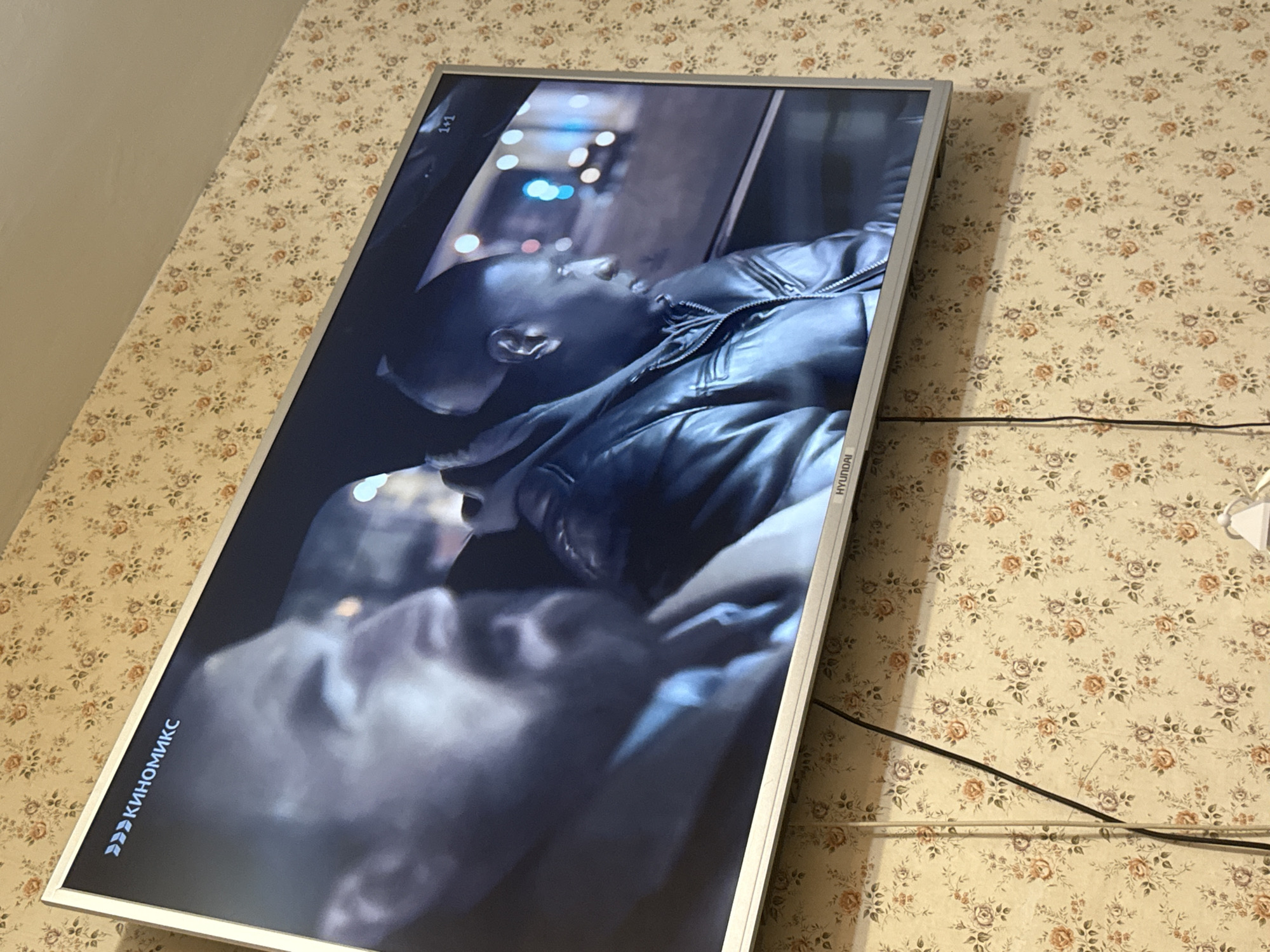 A medium shot shows a television displaying an image of a Black man in a leather jacket, sitting in a vehicle at night. Street lights are visible in the background. The television, a HYUNDAI brand, is mounted on a wall with a floral wallpaper. Below the screen are visible wires and cables. A partial Russian word 'КИНОМИКС' (KINOMIKS) is displayed on the bottom left corner of the screen. The location is Mytishchi, Russia.