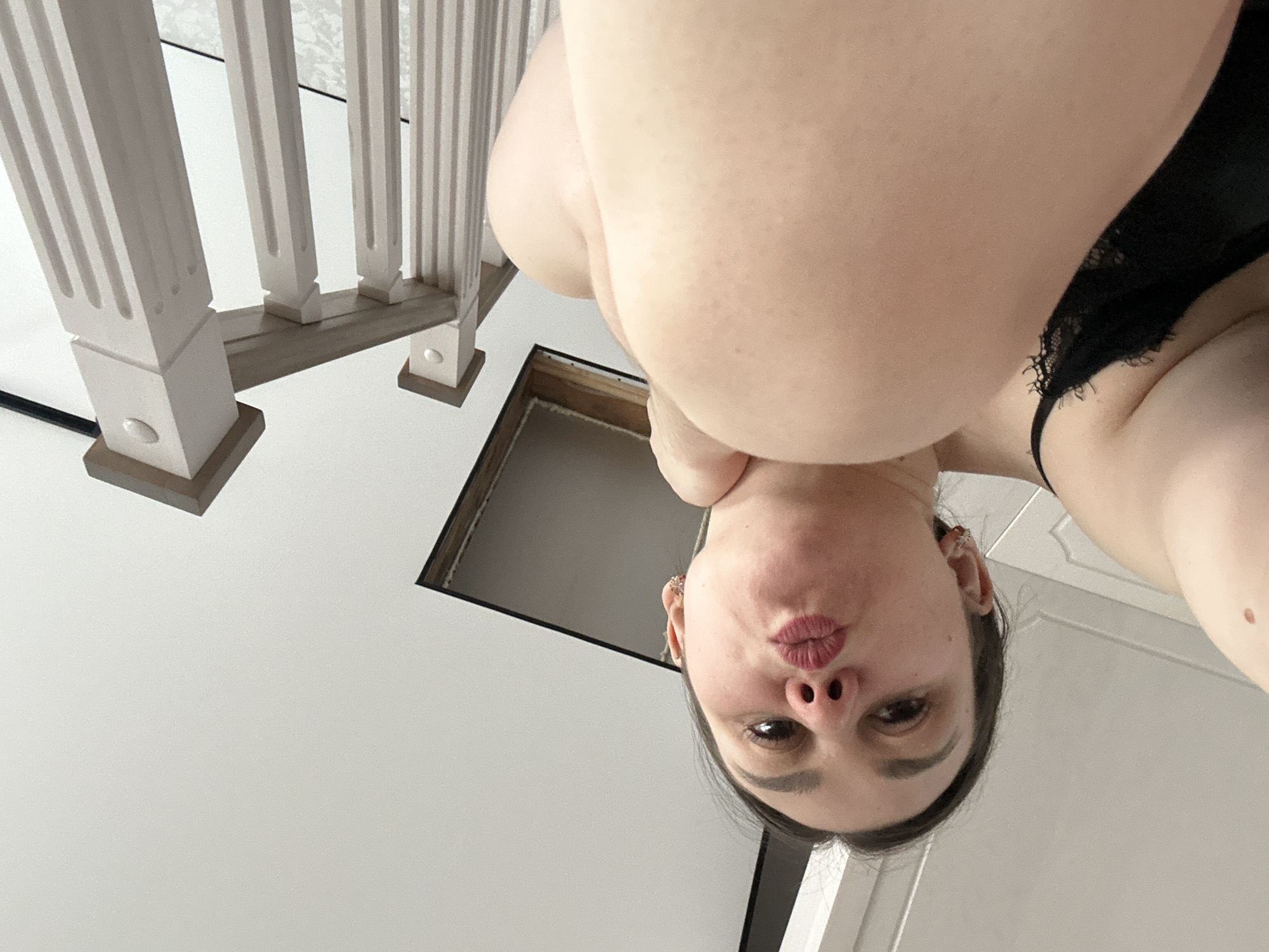 An indoor, low-angle shot captures a female subject looking downwards, her face appearing upside down from the viewer's perspective. She is positioned centrally in the frame, making a playful "puckered lips" expression. Her dark hair is visible around her face, and an ear with a small earring can be seen on her left side. Her upper body is visible, with bare skin on her arms and shoulders, and a glimpse of a black lace garment, possibly a bra or lingerie, on her right side.
To the upper left of the image, a white wooden staircase balustrade with ornate, fluted posts and a darker gray/brown banister is visible. Below the balustrade, an open rectangular void or hatch is seen in what appears to be a floor or ceiling, with dark edges. The background consists predominantly of a plain, bright white surface, likely a ceiling or wall. The lighting is bright and even, characteristic of an indoor setting during daytime. The image conveys a playful or intimate moment, originating from Chaikovsky, Russia. No text is visible in the image.