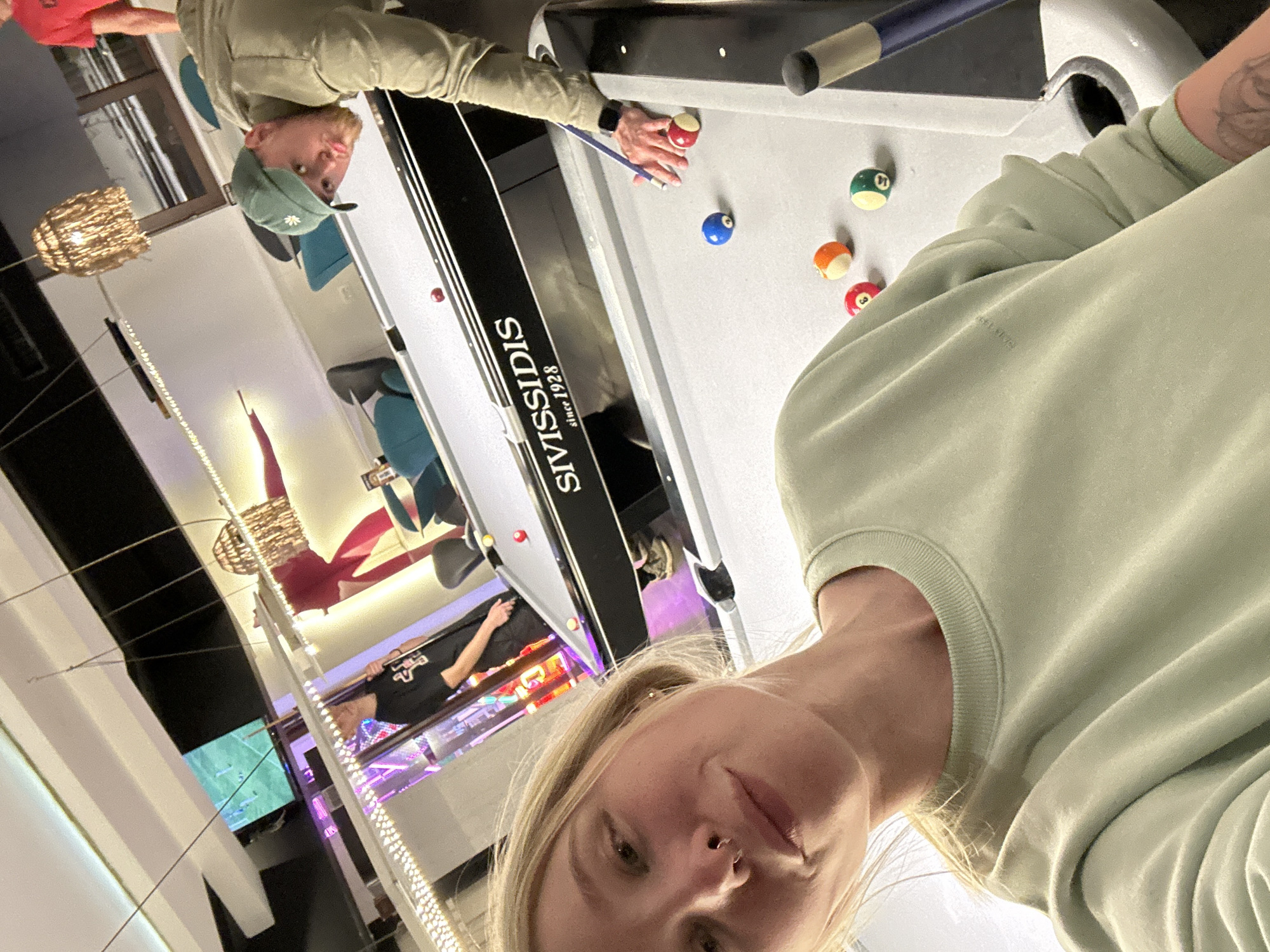 The image shows a selfie taken in a recreational area, likely a bar or entertainment venue, in the city of Budva, Montenegro. A woman with blonde hair and a nose piercing is in the foreground, wearing a light green sweatshirt. Behind her is a billiard table with a brand name "SIVISSIDIS since 1928" visible on its side. Two other individuals are visible around the pool table: one wearing a green cap aiming with a pool cue and another person in the background wearing a black shirt with a red logo. Several billiard balls are scattered on the table. Beyond the pool table and the people, there are decorative lights and a television screen showing a soccer game, further indicating the nature of the venue. The overall lighting of the place is bright.