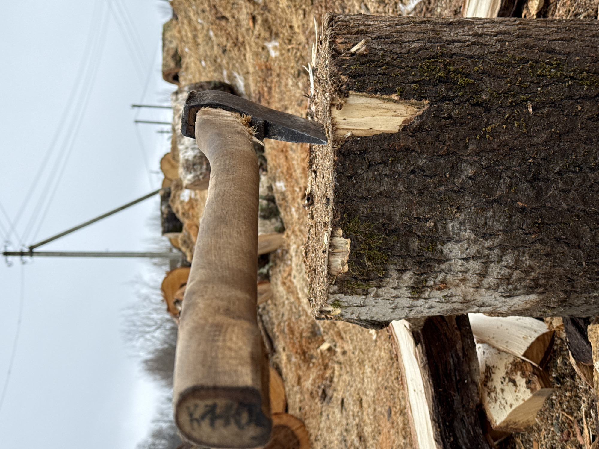 The image depicts a wood splitting scene, likely near Nizhnyaya Chermoda, Russia, although this cannot be definitively confirmed from the image alone. An axe is embedded in a partially split log. The axe has a wooden handle and a dark-colored metal head. The log is roughly cylindrical with bark present. The bark appears dark brown with some green moss. The split shows light-colored wood. Surrounding the log are other pieces of split wood. The background shows a wooded area, potentially with other logs piled up. In the far background, electrical lines and poles can be seen against a grey sky, suggesting a rural or semi-rural setting.