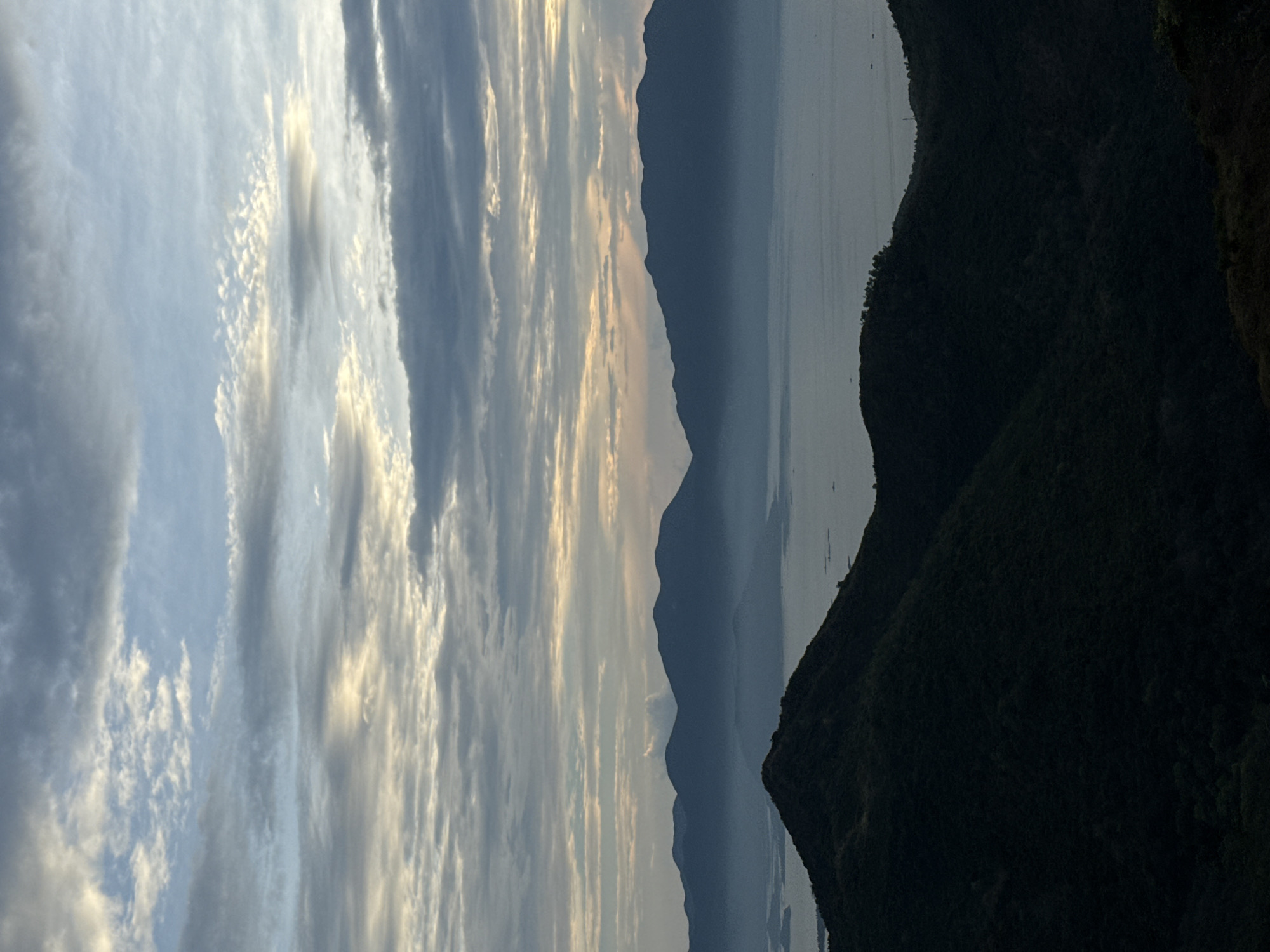 The image shows a landscape view, likely at dawn or dusk, with multiple layers. The sky is filled with striated clouds, ranging from light blue to gray with hints of gold. Below the sky are a series of mountain silhouettes receding into the distance. Between the mountains, a body of water is visible. The foreground features a dark, heavily vegetated mountain range. The image could be taken in or near Bac Nha Trang Ward, Vietnam given the landscape features. There are no visible people or specific actions.