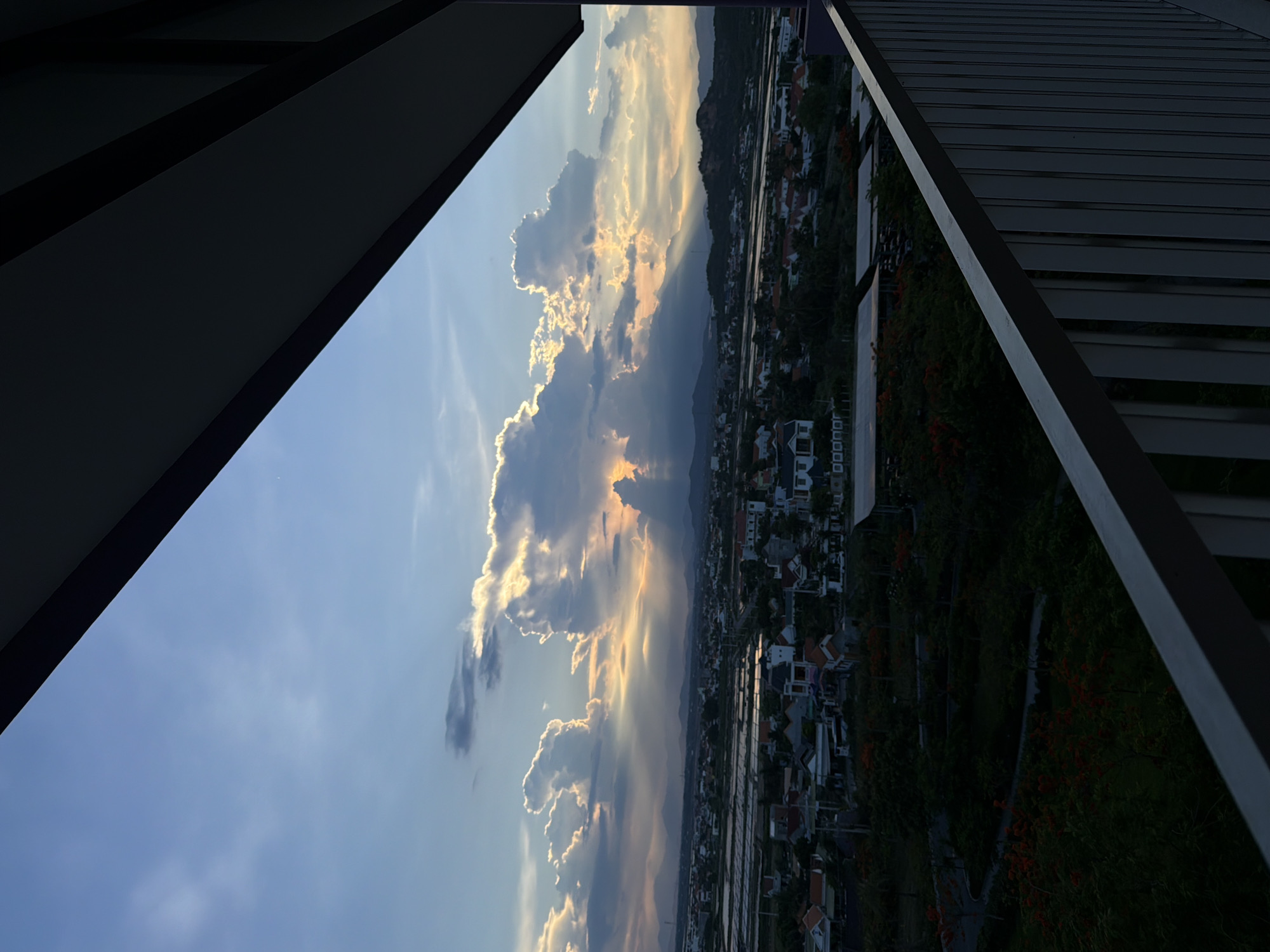 The image is an outdoor scene captured from a high vantage point, possibly a balcony, overlooking a landscape at dusk. The foreground is dominated by a dark, angled structure, likely a balcony railing and ceiling, which frames the view. Below this, a town or village is spread out, characterized by numerous houses with tiled roofs, interspersed with greenery. In the middle ground, there appears to be agricultural land or water bodies, possibly rice paddies, reflecting the sky.
The sky is a prominent feature, filled with dramatic clouds illuminated by the setting sun. Rays of light pierce through the clouds, casting shadows on the landscape below. The colors in the sky range from a pale blue to warm oranges and yellows, indicating the golden hour. In the distance, mountains form a silhouetted horizon. The overall atmosphere is serene and picturesque, typical of a sunset view in a rural or semi-urban setting.
While the prompt specifies "Dong Ninh Hoa Ward, Vietnam," this detail cannot be visually confirmed from the image alone. The scene depicts a generalized Vietnamese landscape with its characteristic architecture and vegetation. There are no people or specific activities visible in the frame. The focus is on the natural light and the interplay of clouds and landscape.