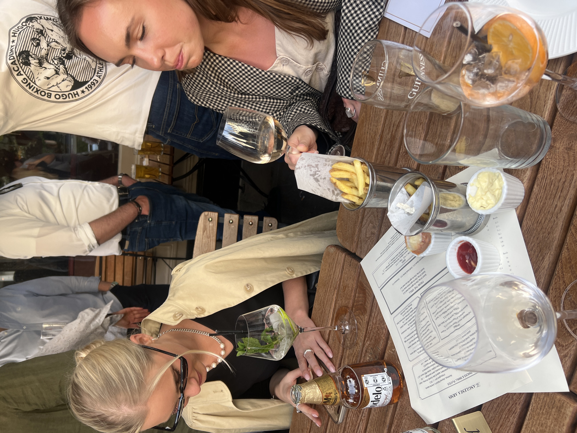 This image captures a casual outdoor gathering, likely at a pub or restaurant patio in London, United Kingdom, given the "The ANGLESEA ARMS" menu visible. The scene is set around a wooden table during what appears to be daytime, possibly late afternoon, with natural light.
Three women are prominent. In the foreground, one woman with blonde hair, wearing glasses and a light trench coat over a black top, holds a Modelo beer bottle and a glass containing a clear drink with mint and ice. She is also wearing a pearl necklace and a silver chain. Beside her, another woman with brown hair, dressed in a plaid jacket over a light top and blue jeans, rests her head on what appears to be someone's arm or shoulder, eyes closed, suggesting comfort or weariness. In the background, a man's torso is visible, wearing a white t-shirt featuring a "HUGO BOXING ACADEMY 1993" logo. Other patrons are partially visible in the blurred background.
The wooden table is laden with various items: several glasses, some empty, one containing white wine, and another with an orange slice and ice. Two metal holders filled with french fries are present, alongside small white ramekins containing mayonnaise and ketchup. Menus from "The ANGLESEA ARMS" are spread on the table, detailing "SNACKS & SMALL PLATES" and "FROM THE GRILL" options, including "Scotch 35 day 10oc dry aged rump sta 31.5", "Chips 6.5", and "MODELO" on the beer bottle. A "GUINNESS" logo is faintly visible on one glass. The overall atmosphere seems relaxed and social, with people enjoying food and drinks.