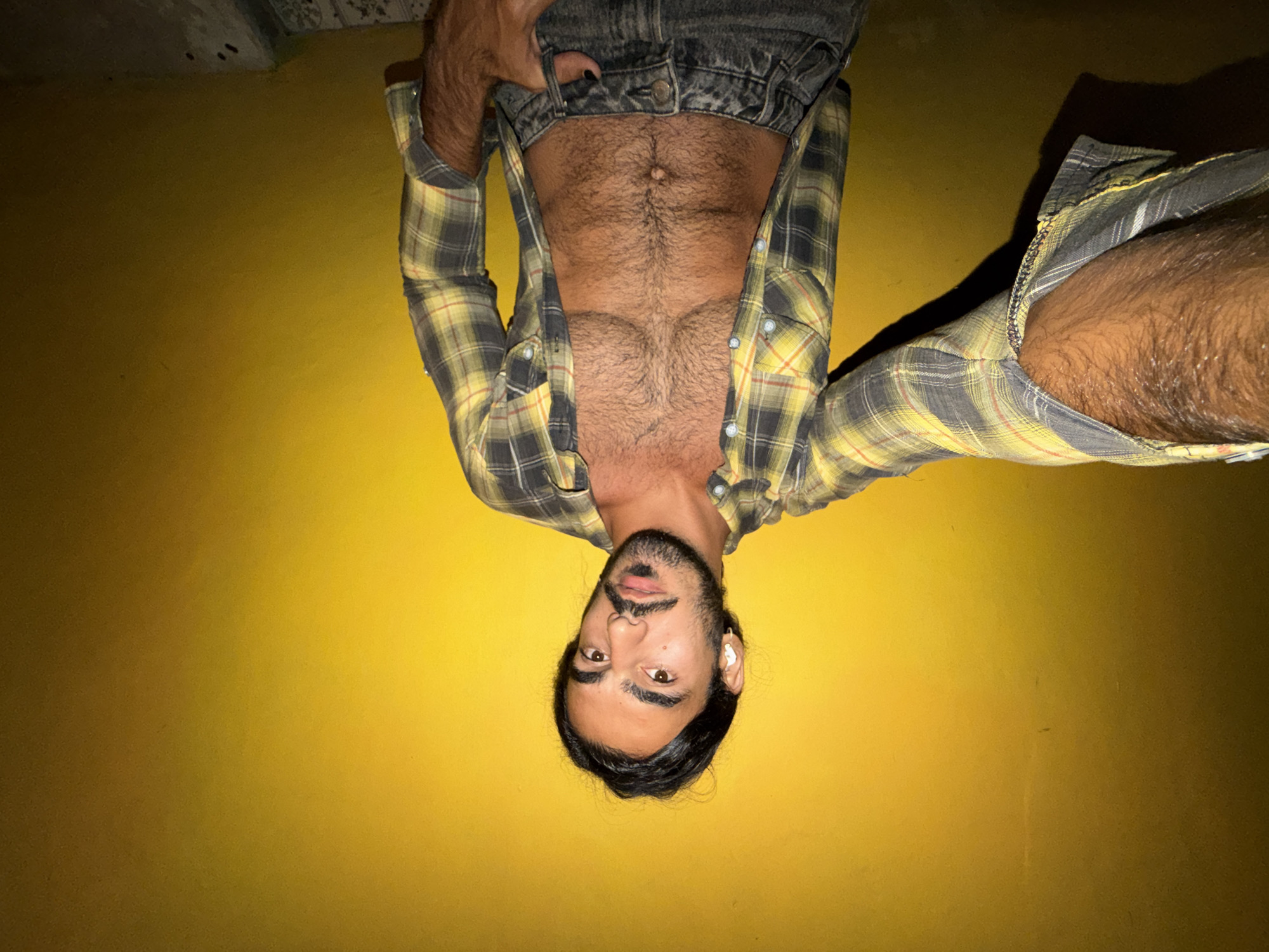 A man with dark hair and a beard, wearing a plaid shirt unbuttoned to reveal his chest, stands in front of a bright yellow wall. He has an earbud in his left ear and is holding the camera for a selfie. His right hand is in his pocket. The lower half of his body is visible, wearing dark jeans. The background appears to be a wall with some lighting effects, possibly in an outdoor urban setting. The scene could be in the city of North Kuta, Indonesia, given the warm lighting and texture of the wall.