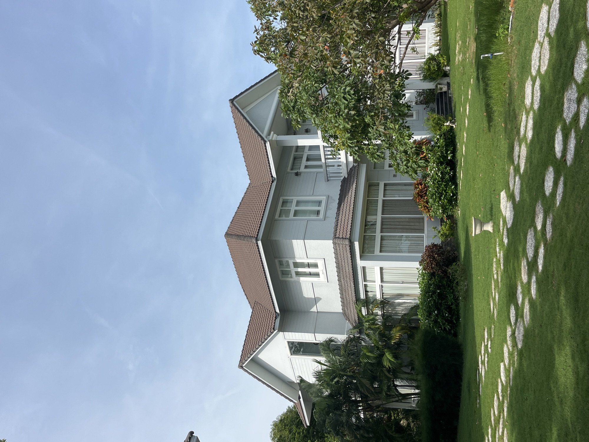 The image depicts a modern, two-story house with a brown tiled roof, light gray siding, and white-trimmed windows. A balcony with white railings is visible on the upper floor. The house is surrounded by a lush green lawn, with a winding path of flat, irregularly shaped stones leading across it. Several trees and various types of foliage, including palm trees, are planted around the property. The sky is clear blue with a few wispy clouds. The scene appears to be a residential property in a tropical or subtropical climate. The location is identified as Phu Thuy Ward, Vietnam, suggesting a suburban or semi-rural setting. There are no people visible in the image.