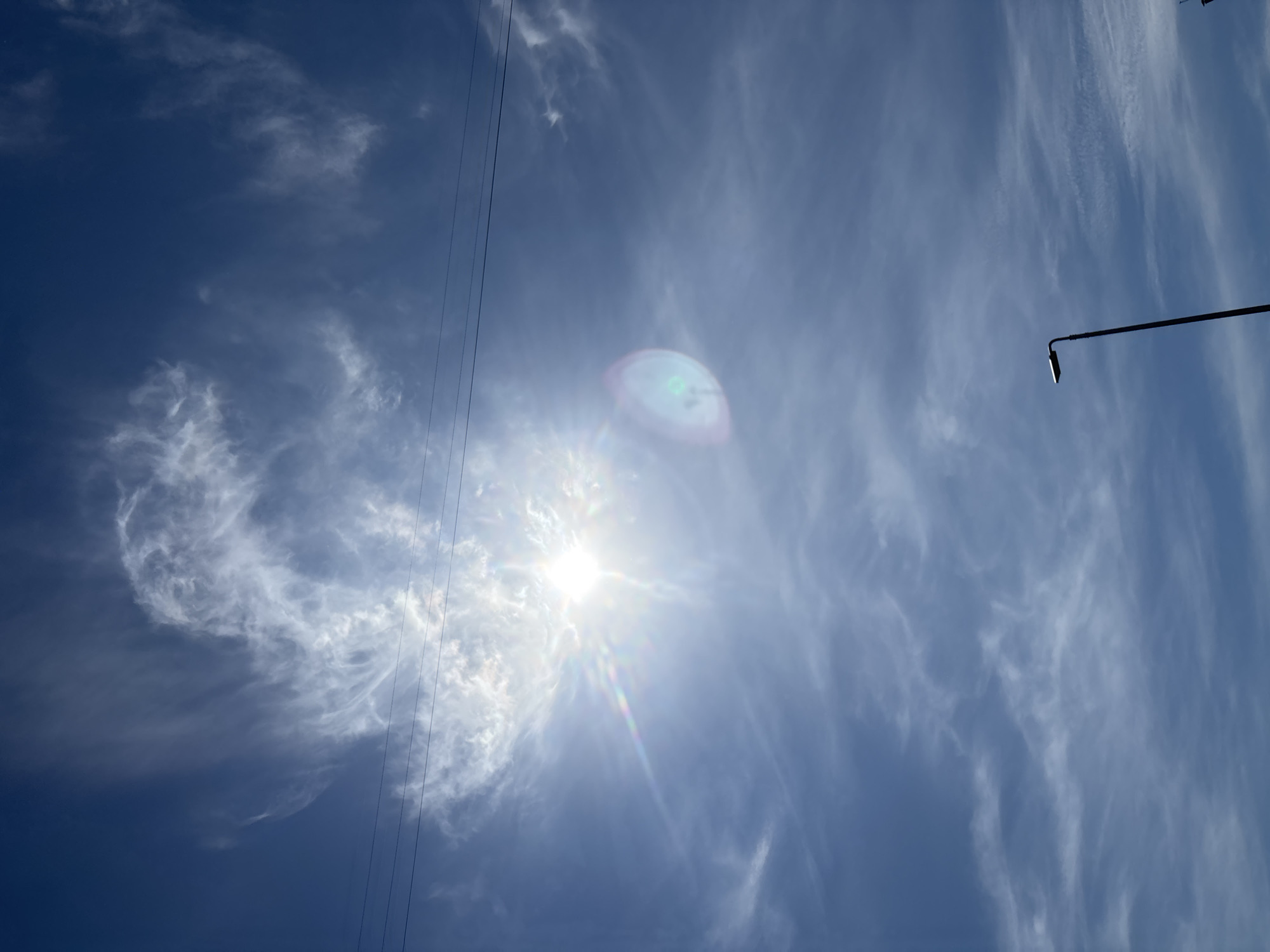 The image shows a view of the sky in Chaikovsky, Russia. The sky is blue with streaky white clouds. The sun is visible, creating a bright glare and lens flare. Overhead power lines are visible on the left side of the image. A lamppost extends into the sky from the right side of the image.