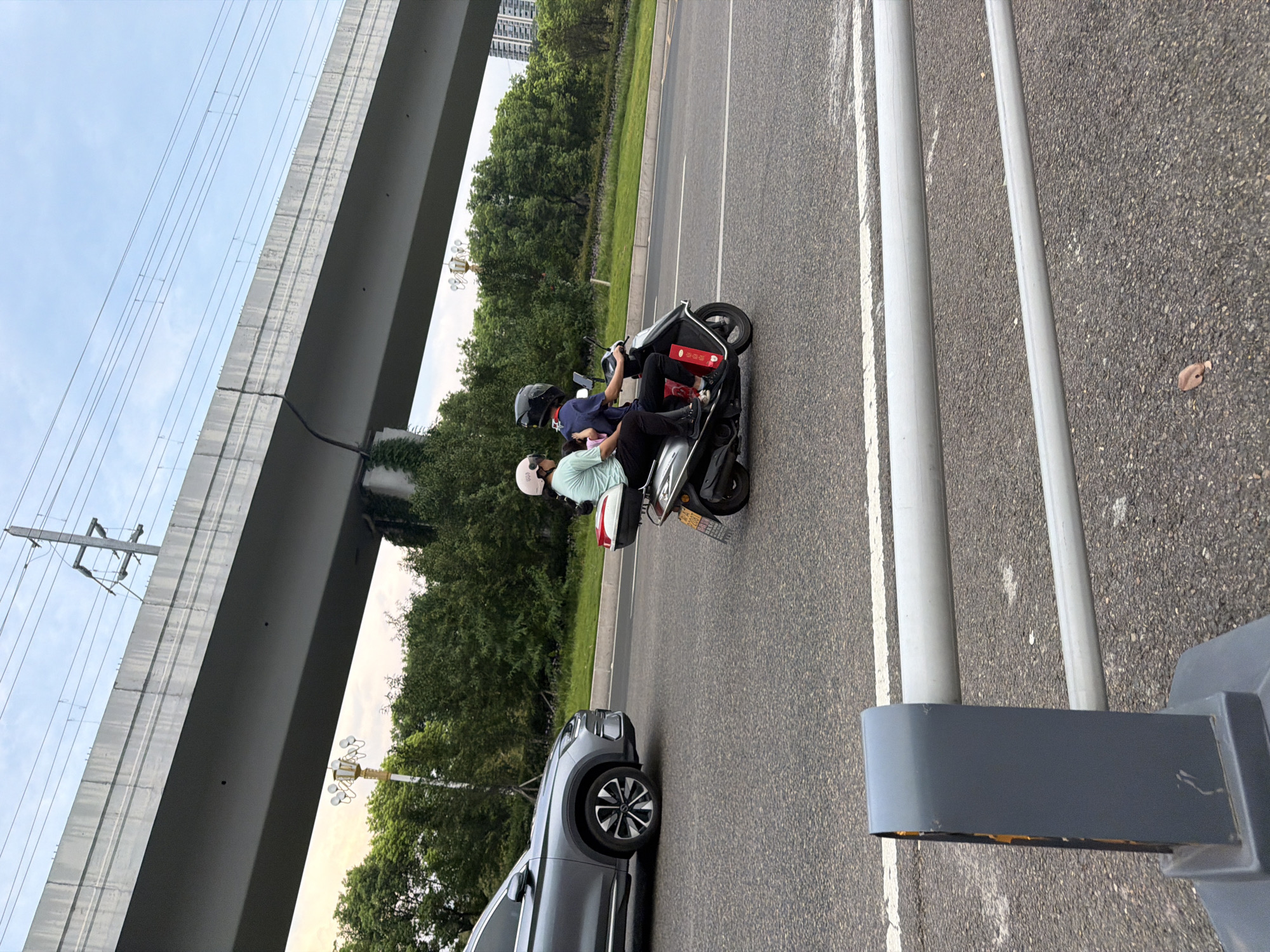 In Jiangjin, China, a gray SUV drives on an asphalt road under an elevated highway structure. To the right of the SUV, two people ride on a silver scooter. The person in front wears a light blue t-shirt and a pink helmet, while the person behind wears a dark blue shirt and a black helmet. The scooter has a red and white storage box on the back. Trees and a lamppost are visible behind the road, and distant buildings are in the background. The sky is cloudy. In the foreground, a gray barrier with silver pipes runs across the image.