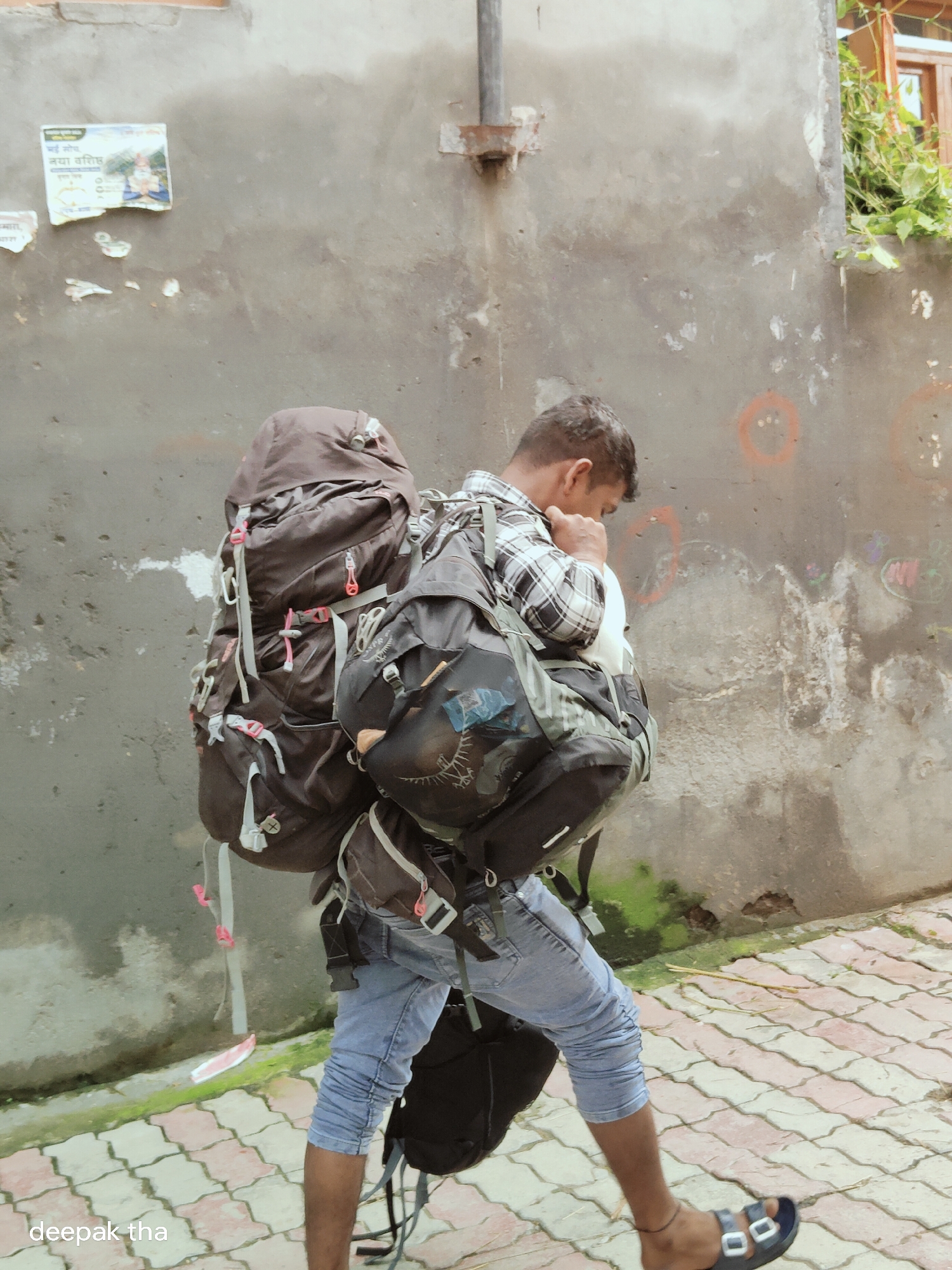 A young man wearing a plaid shirt and blue jeans walks to the right, carrying a large brown backpack on his back and another black backpack in front. He is in the village of Vashisht, India, near the Tibetan market. The man is walking on a tiled path beside a rough, grey concrete wall. The wall has some peeling paint, graffiti, and posters. Green foliage is visible growing near a window on the upper right side of the wall.