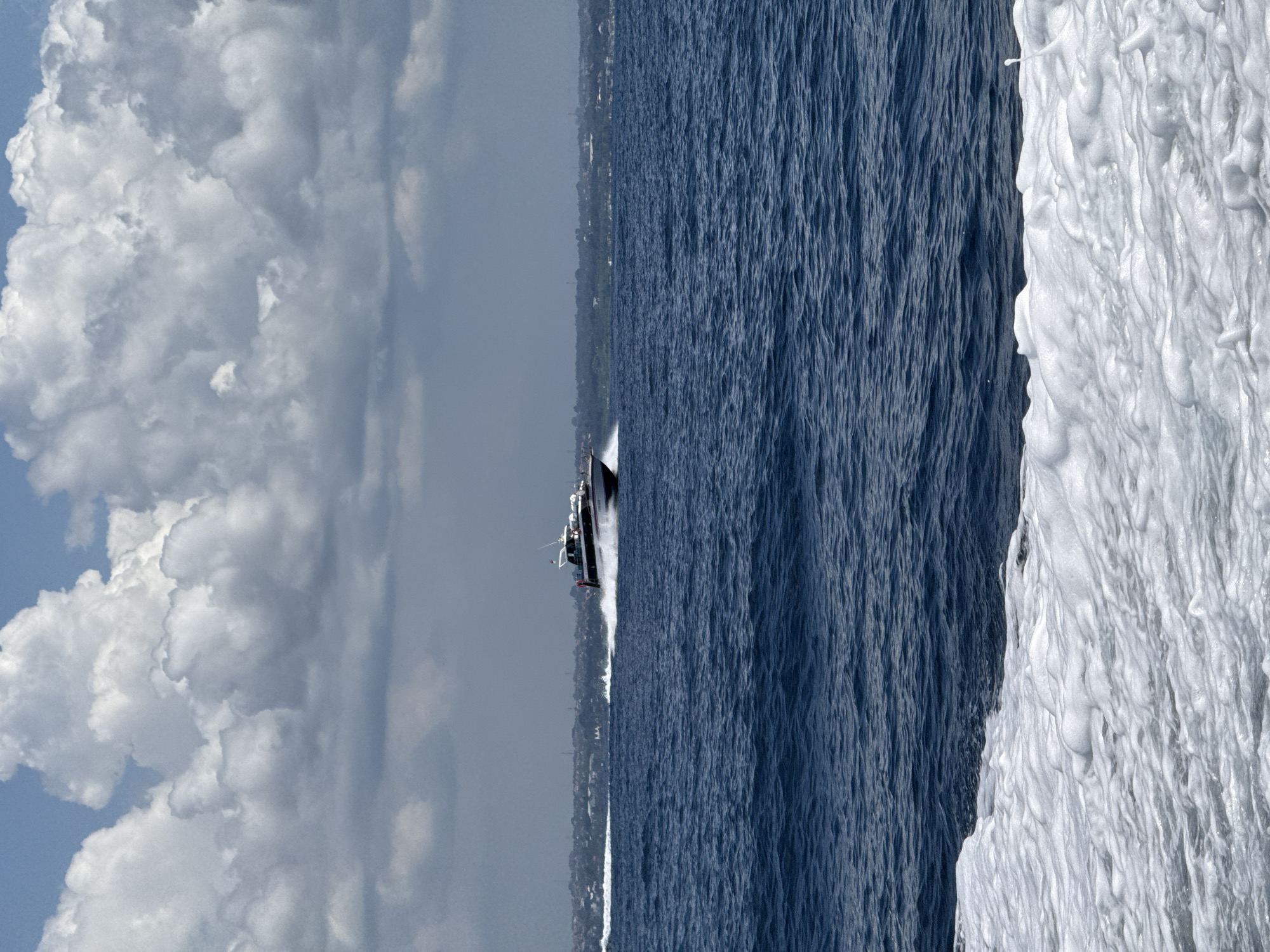 The image shows a body of water with a boat moving through it. The boat is in the middle of the frame, with a trail of white water behind it. The sky is on the left side of the image and is filled with white, puffy clouds. The water is a deep blue color and has white foam on the right side of the frame. In the distance, there is a strip of land covered in trees. The location is Unknown, Indonesia.