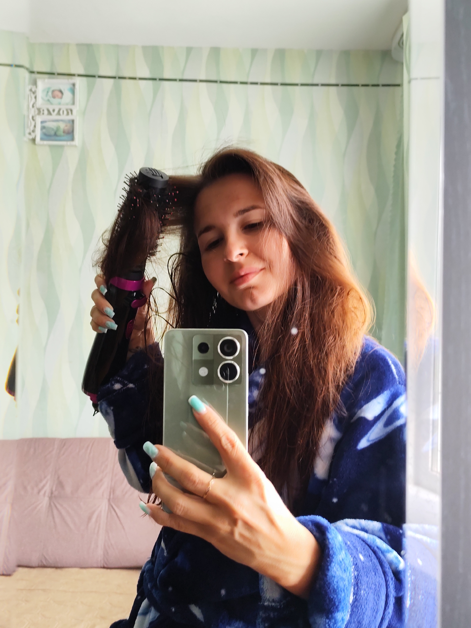 A woman is taking a mirror selfie while styling her hair. She holds a smartphone in her right hand with light blue painted nails and a ring on her finger. In her left hand, she holds a black and pink hot air brush, using it to style her long, reddish-brown hair. The woman wears a blue robe with a white star pattern. The background shows a light green wallpaper with white wavy lines. Above her, there's a decorative picture frame with baby photos and Cyrillic text. Behind her, a portion of a light pink couch is visible. To the right, a white window frame is apparent. There is no indication that the image was taken in the city of Фоки, Russia.