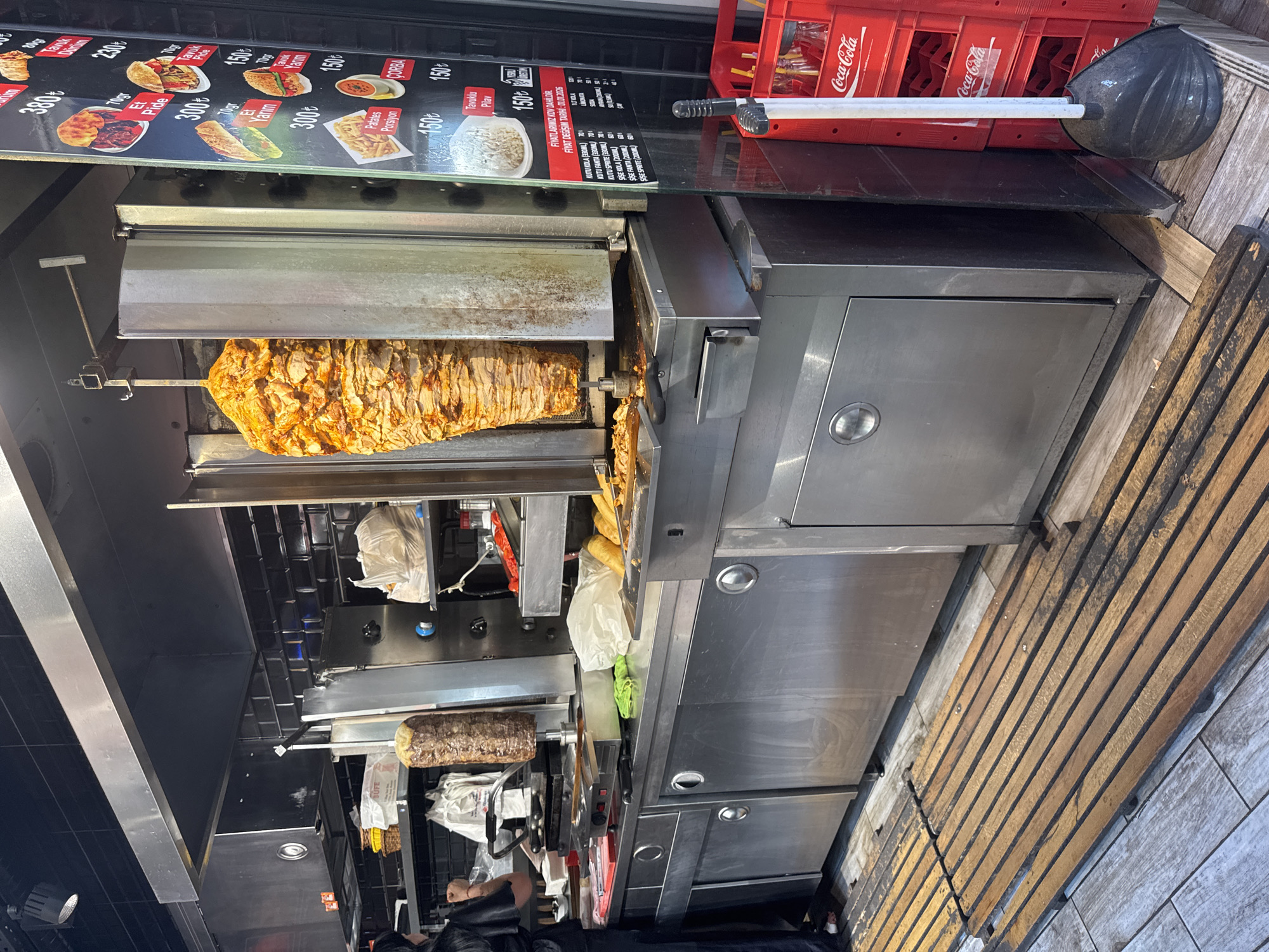 The image shows an outdoor food stall in the Gungoren district of Istanbul, Turkey, specializing in doner kebab. A large vertical spit of seasoned meat, likely chicken, is slowly rotating and being shaved off by an unseen chef. In the foreground, a wooden slatted platform covers the ground in front of the stall. Behind the meat spit, stainless steel counters and equipment are visible, including a grill and storage units. A menu board is displayed on the right side of the image, listing prices in Turkish Lira. A stack of red Coca-Cola crates is also present, along with a dustpan and brush. A person, partially visible on the left, appears to be working behind the counter. The overall scene depicts a busy food service environment with typical street food elements.