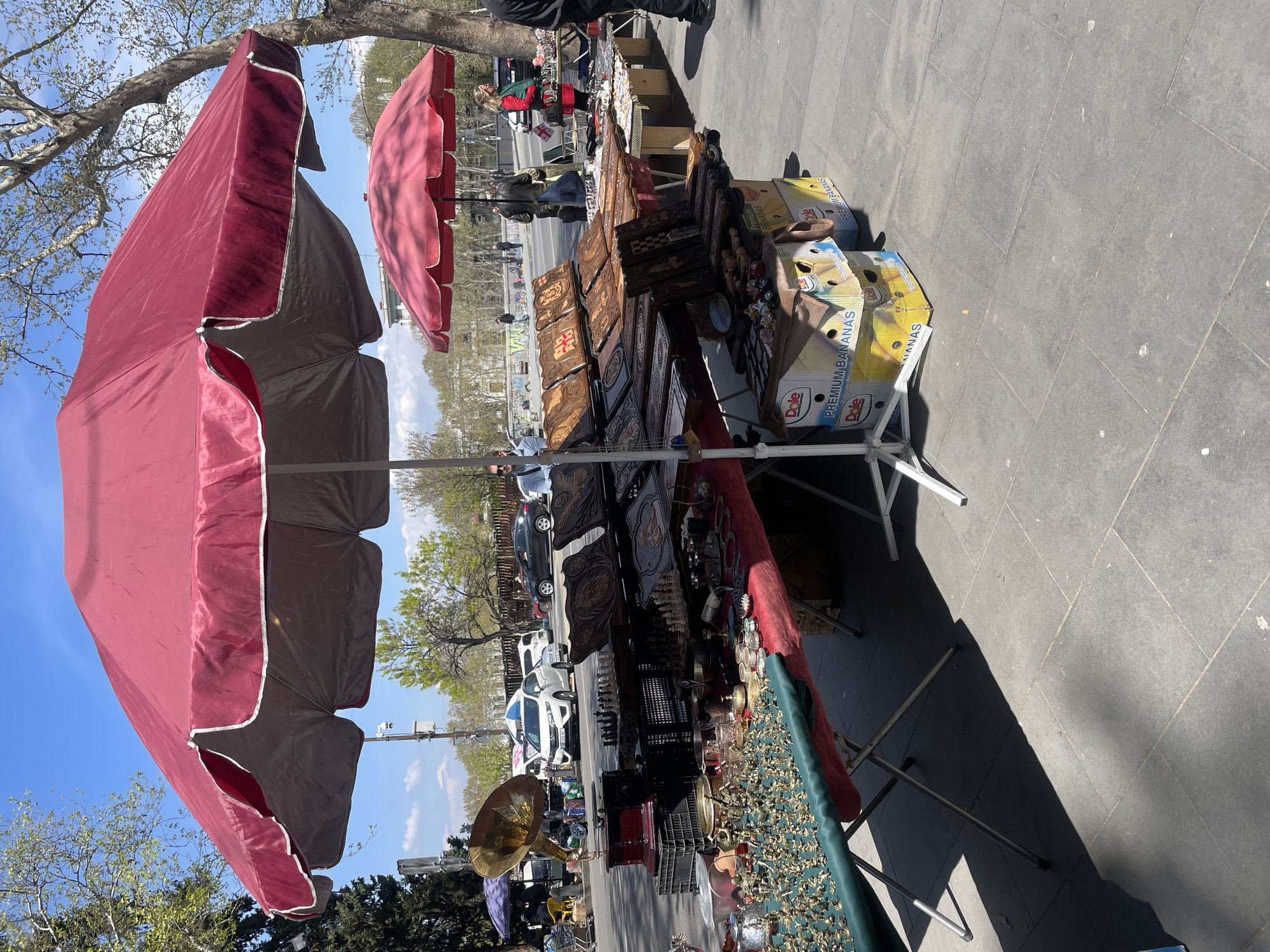 An outdoor street market scene unfolds under a clear blue sky, illuminated by bright sunlight which casts distinct shadows. Two large, open red-maroon umbrellas provide shade over the vendor stalls on a paved sidewalk.
The primary market stall in the foreground displays a diverse array of items for sale. Prominently featured are numerous intricately carved wooden boxes and panels, many adorned with distinct geometric and cross patterns. Alongside these, smaller metal trinkets, possibly jewelry or decorative figurines, are laid out on a green-covered surface. A large, polished brass horn also stands out among the merchandise. The stall's display incorporates cardboard boxes labeled "Dole PREMIUM BANANAS" as makeshift risers and storage.
In the background, a street is visible with several cars, both parked and moving. The street is flanked by trees with early spring foliage. Several people appear as blurry figures, some walking along the street, others seemingly browsing at distant stalls. One person in a red jacket is more clearly visible near a stall further down the market row. The scene captures a bustling, sunny day at what appears to be a typical urban street market or flea market, consistent with a setting like Tbilisi, Georgia.