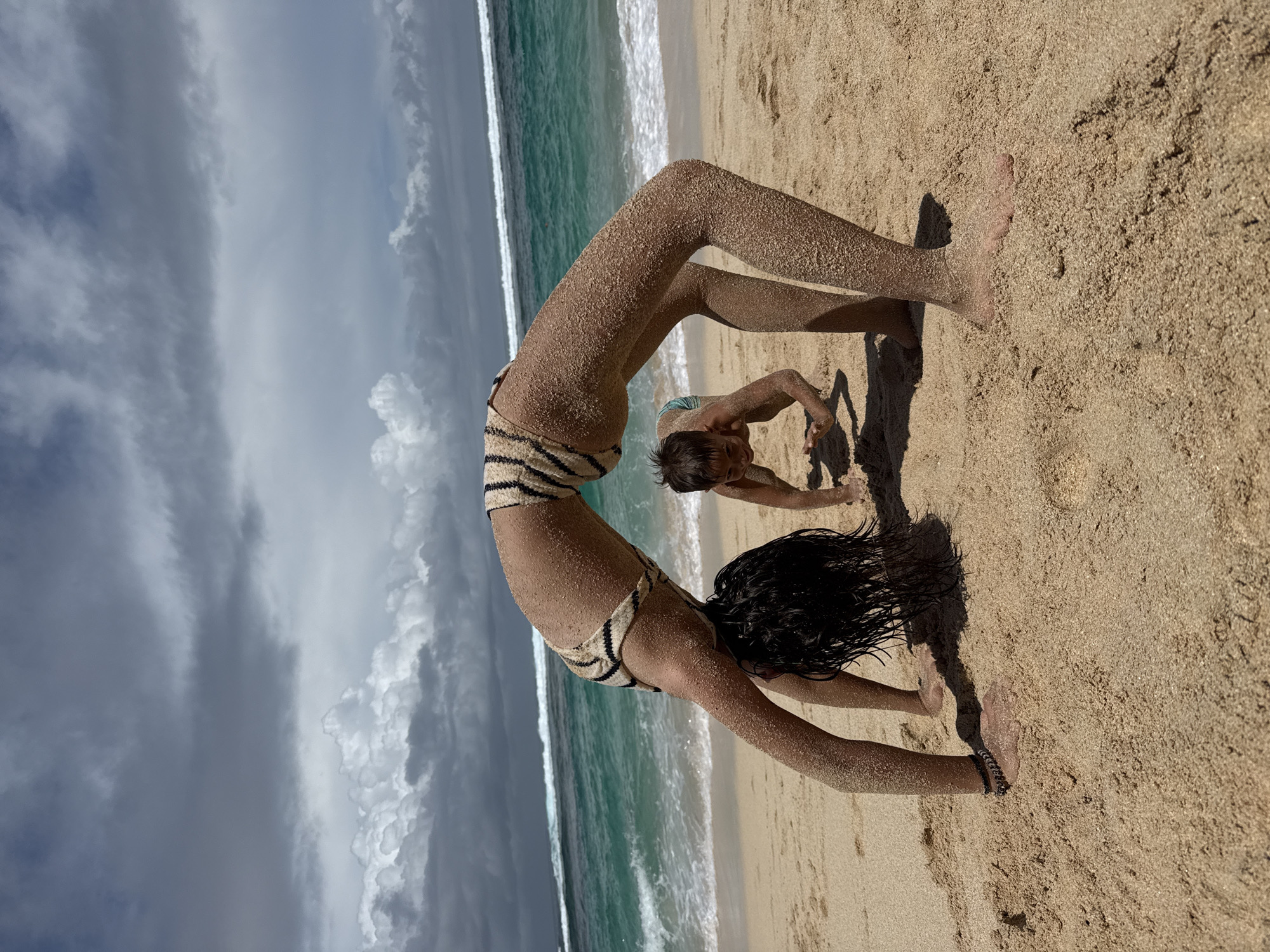 An outdoor, eye-level shot captures two individuals on a sandy beach in Kuta Selatan, Indonesia. The image is rotated 90 degrees to the left, showing the horizon vertically.
In the foreground, a person with dark, wet hair performs a backbend (wheel pose) on the sand. They are covered in sand from head to toe, wearing a striped (cream and dark brown/black) two-piece swimsuit. Their hands and feet are planted firmly on the sand, supporting their arched body. A dark bracelet is visible on their right wrist.
Behind and slightly to the right of the first person, a younger individual, also heavily coated in sand, is on all fours, looking towards the camera. This person appears to be wearing a light-colored swimsuit.
The setting is a tropical beach. The foreground is dominated by golden sand, which extends to the turquoise ocean in the background. White waves crash near the shore, indicating active surf. The sky is partly cloudy, with a mix of darker gray and lighter white clouds, suggesting a dynamic weather pattern. Shadows cast on the sand indicate daylight, likely mid-morning or afternoon. No visible text is present.