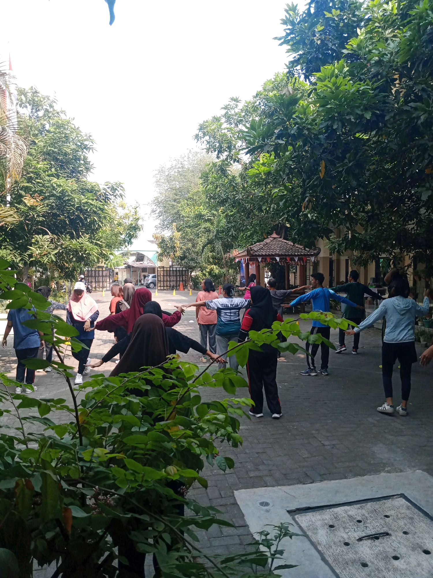 In Surakarta, Indonesia, a group of people is engaged in outdoor physical activity, likely exercise, on a paved courtyard. The setting appears to be a communal space, possibly a schoolyard or community park, surrounded by mature trees. Several individuals, mostly women wearing athletic attire and some with headscarves, are positioned with their arms outstretched, indicating they are performing a coordinated movement. The foreground features lush green foliage, partially obscuring the view of the participants. In the background, a traditional Indonesian gazebo, a building with arched entrances, and a fence are visible, suggesting a cultural or institutional environment. The overall atmosphere is one of active recreation under a bright, possibly sunny sky.