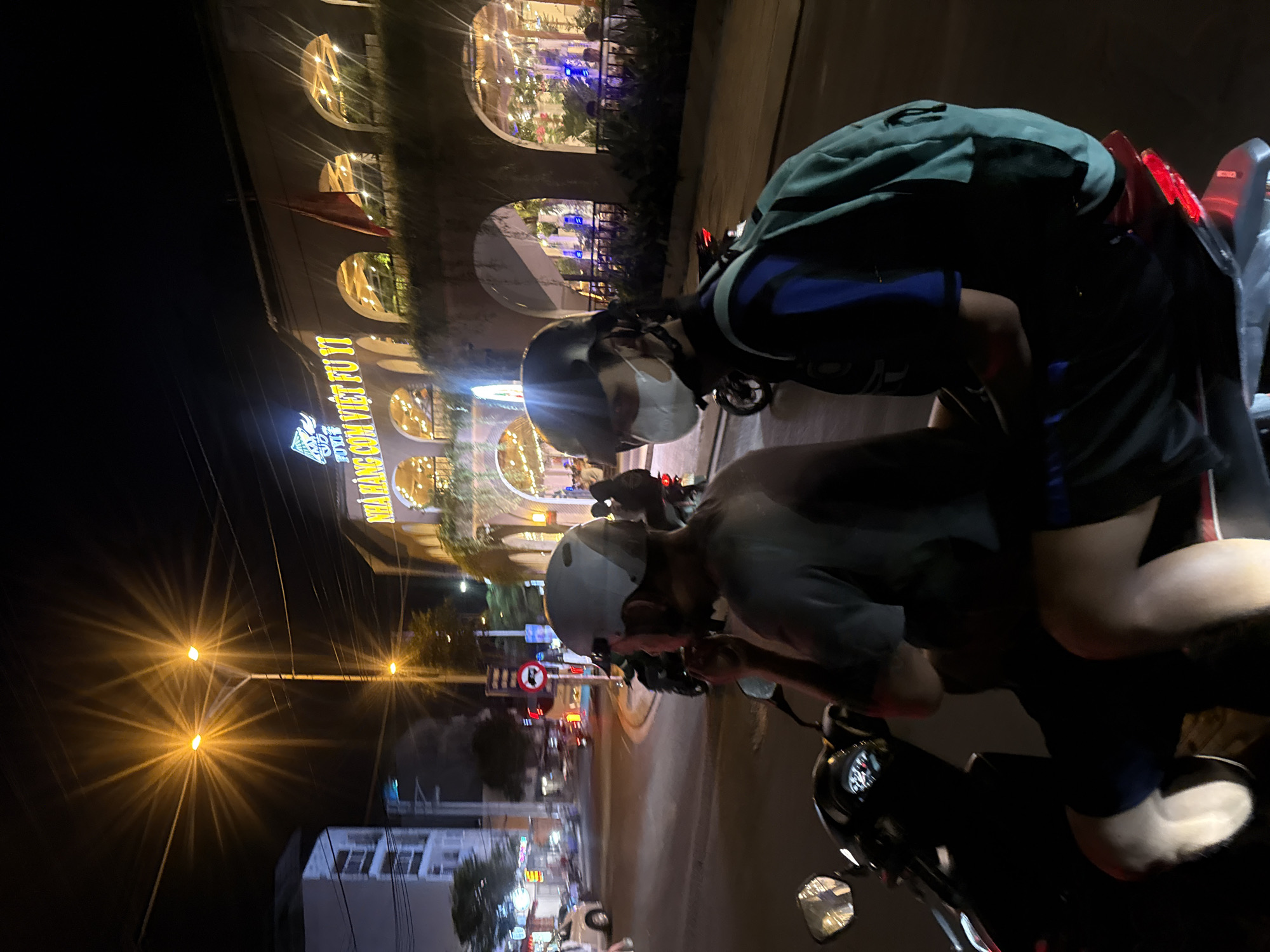 Two people are on a motorbike at night in Bac Nha Trang Ward, Vietnam. The person in front is wearing a helmet and a face mask, and is looking down. The person behind is wearing a green jacket and blue shorts, and is also wearing a helmet. They are stopped next to a building with arched windows lit from within. A sign with Vietnamese text and a logo is visible on the building. Streetlights with bright, starburst-like halos illuminate the scene, casting a warm glow on the road and surrounding area. The overall atmosphere is busy, with some street activity suggested by the lights and traffic in the background. The time of day is clearly night, indicated by the darkness and artificial lighting.