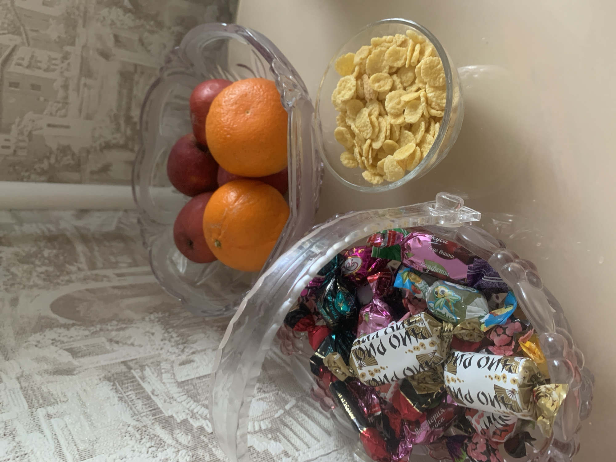 A still-life scene shows three glass bowls arranged on a surface with a patterned design.
The bowl on the left contains several red apples and two oranges.
The bowl in the middle contains yellow cornflakes.
The bowl on the right contains candies of different colors and wrappings. The labels on some of the candies show Cyrillic script.
The background is a light beige wall.
The location is likely Chaikovsky, Russia, given the Cyrillic writing.
